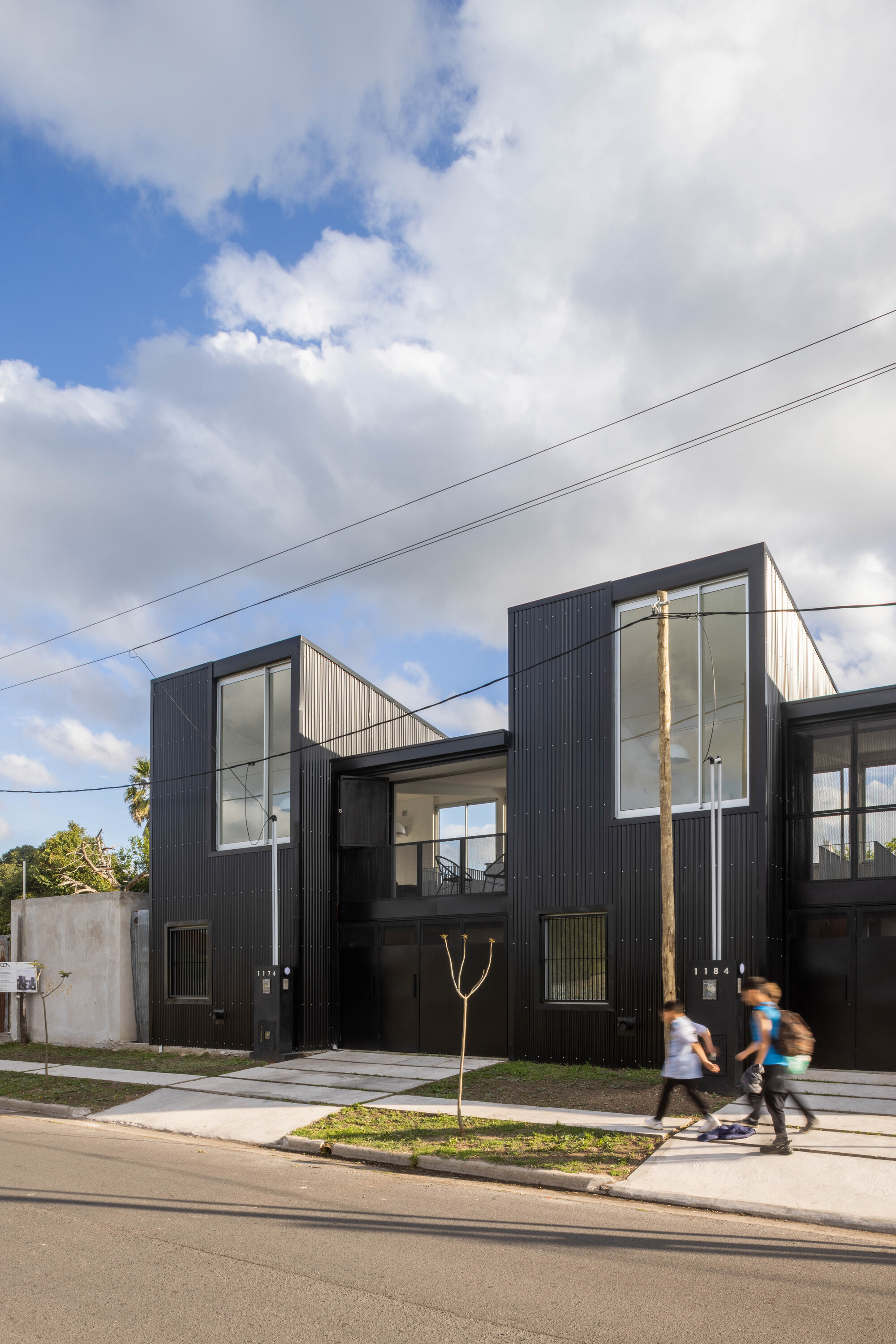 Street view of the black corrugated metal facade with glazed volumes and two people cycling past on the sidewalk