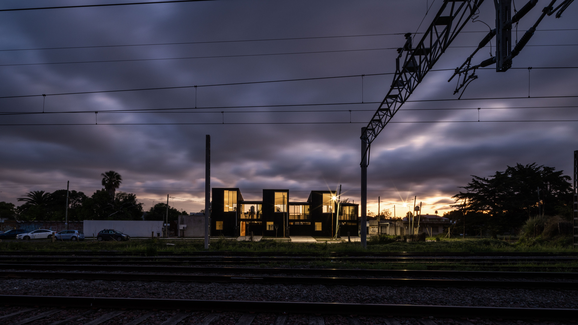 View across railway tracks of the illuminated glass facade at dusk with overhead catenary wires