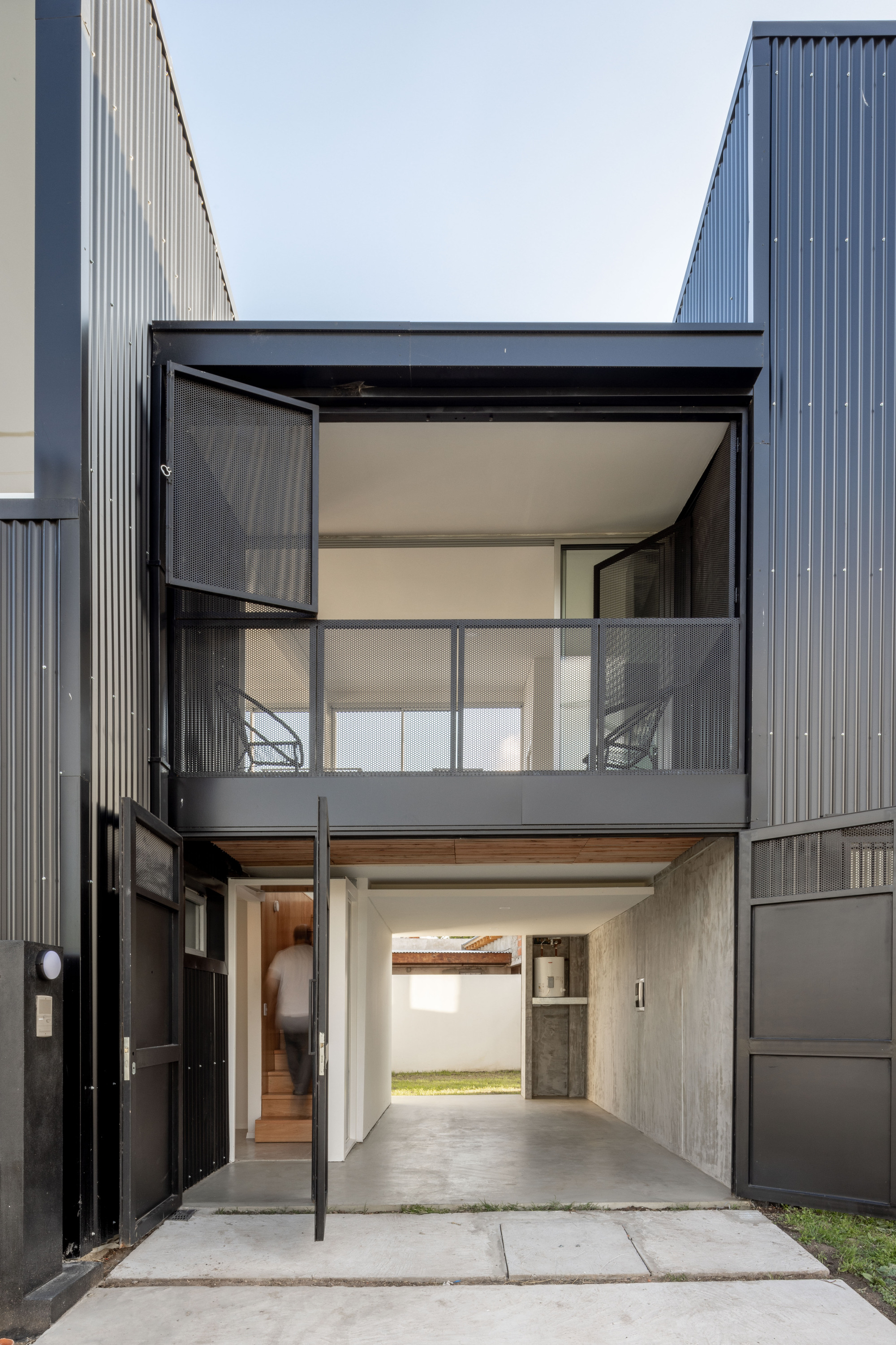 Entry courtyard with pivoting metal door, concrete paving and an upper balcony with mesh railing