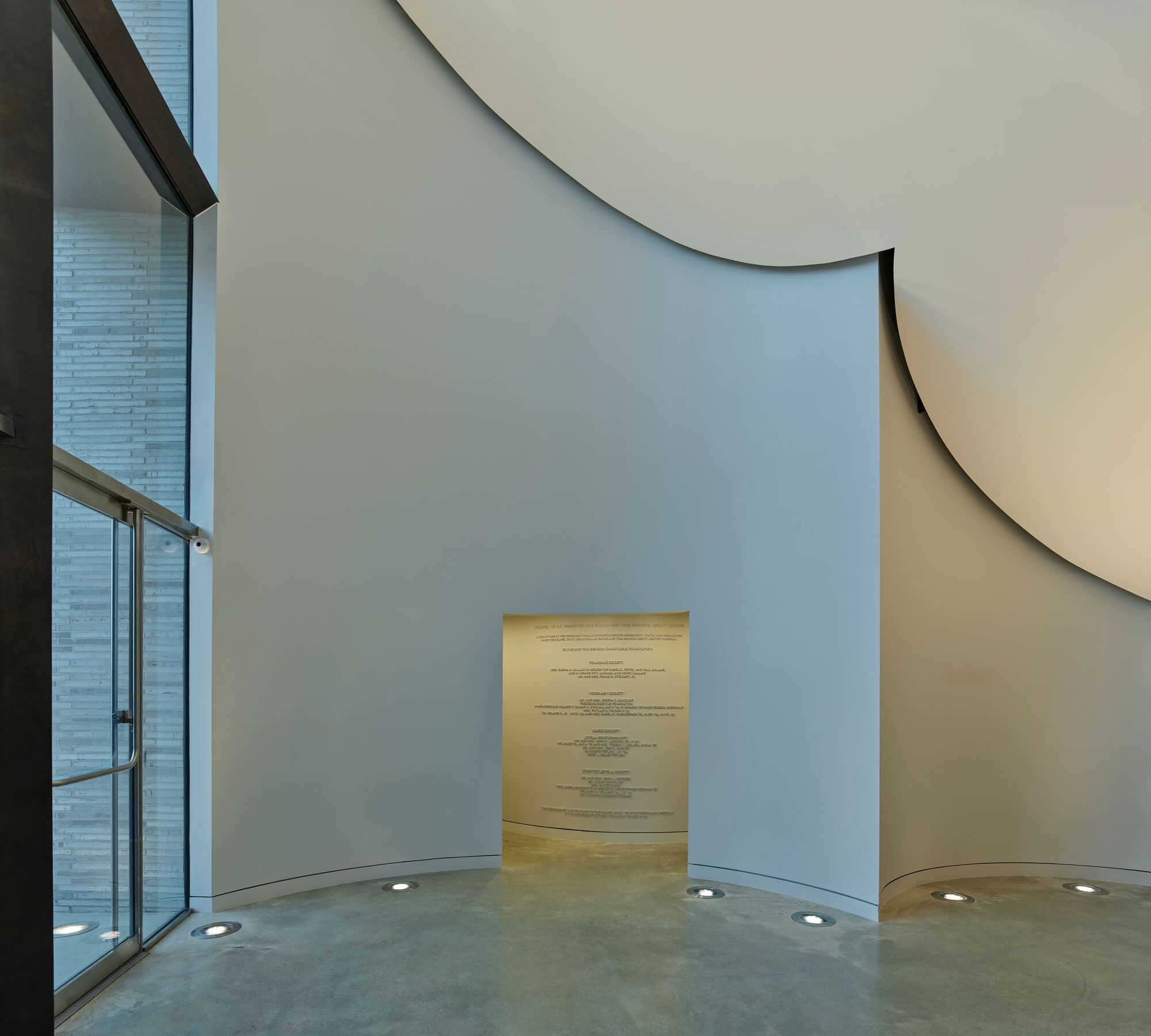 Curved interior wall with narrow illuminated doorway and floor-recessed uplights beside glazed facade