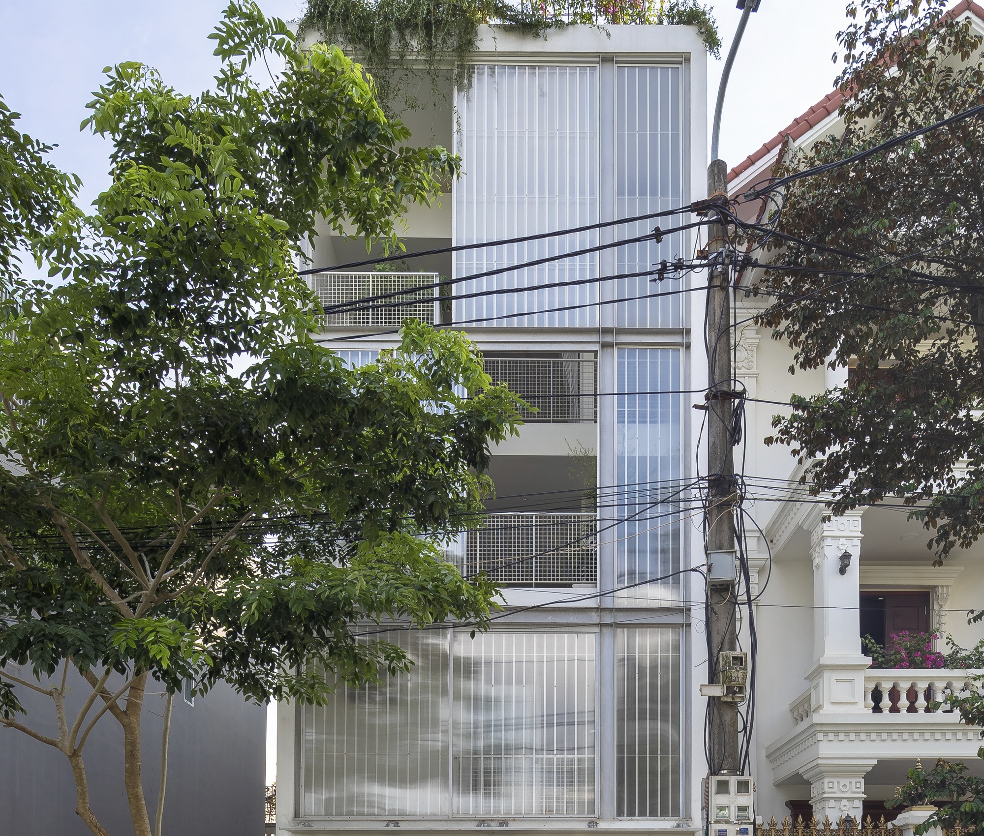 Street view of the vertical corrugated facade with perforated metal balconies framed by neighboring trees