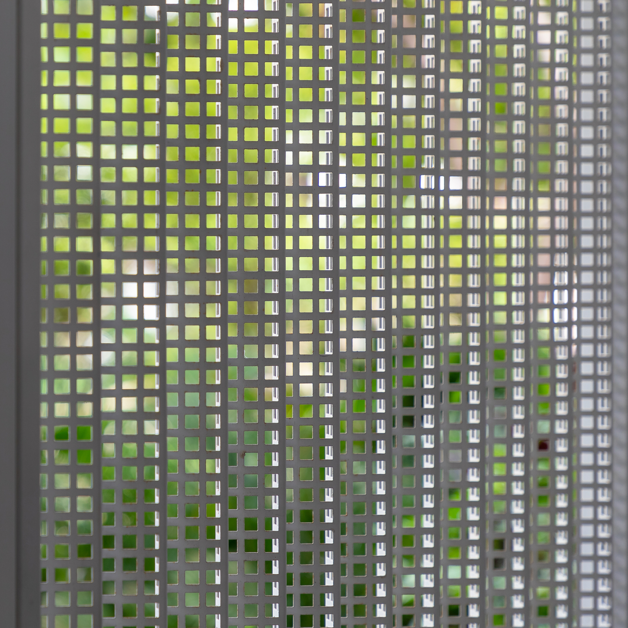 Close-up of perforated metal screen filtering greenery and daylight through square openings