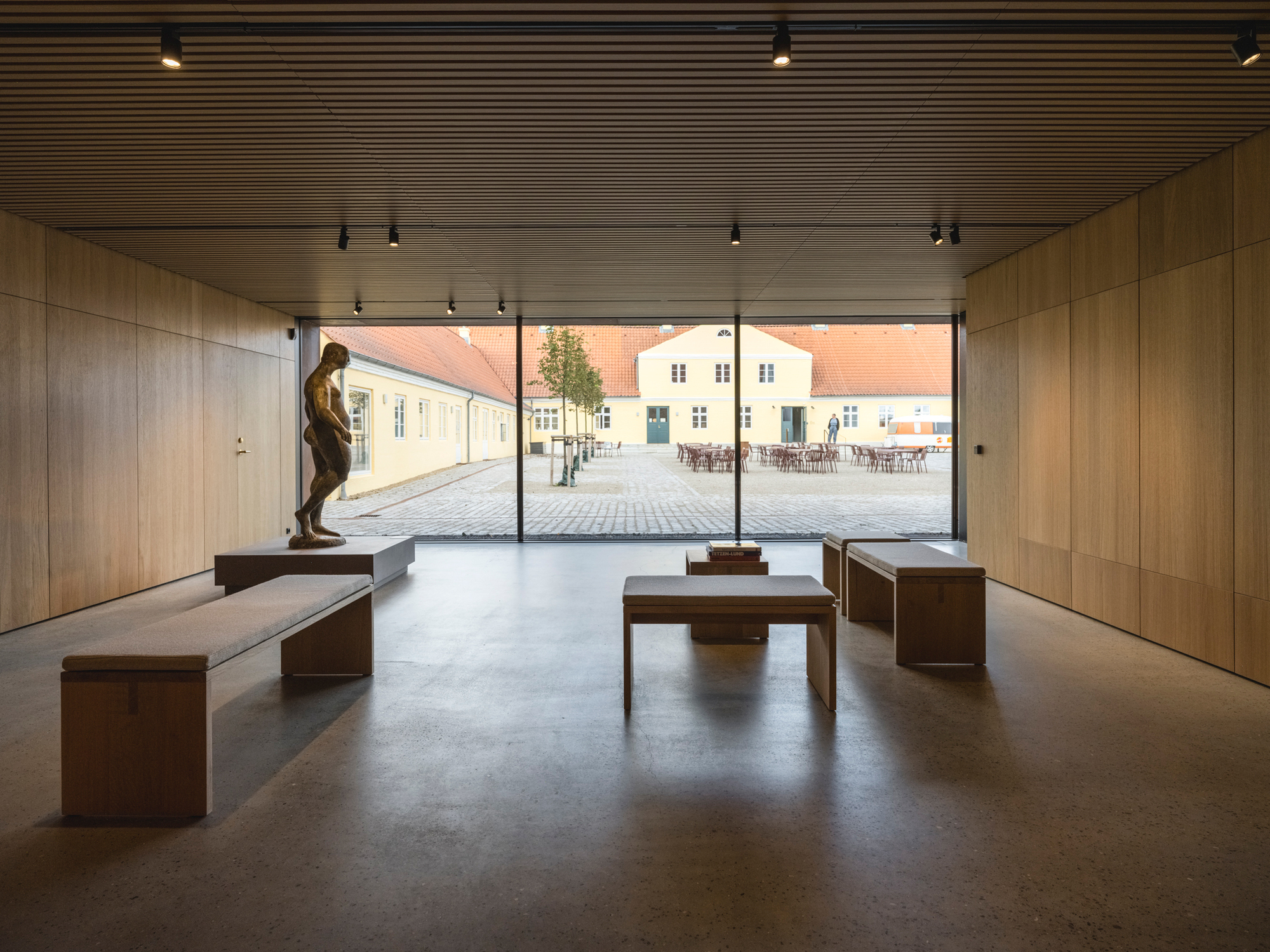 Interior gallery with timber-slatted ceiling, Corten steel benches, and glazed opening framing the courtyard beyond