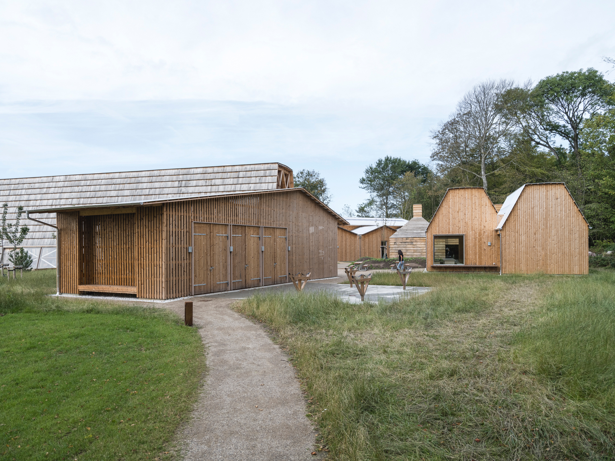 Collection of timber-clad pavilions with varied gabled roofs arranged around a gravel path and meadow planting
