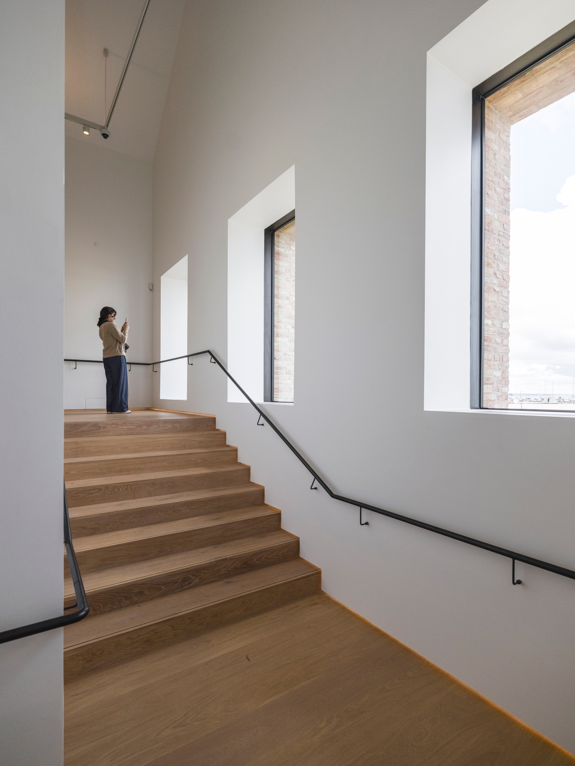 Wide timber staircase with metal handrail ascending past tall windows in white gallery space