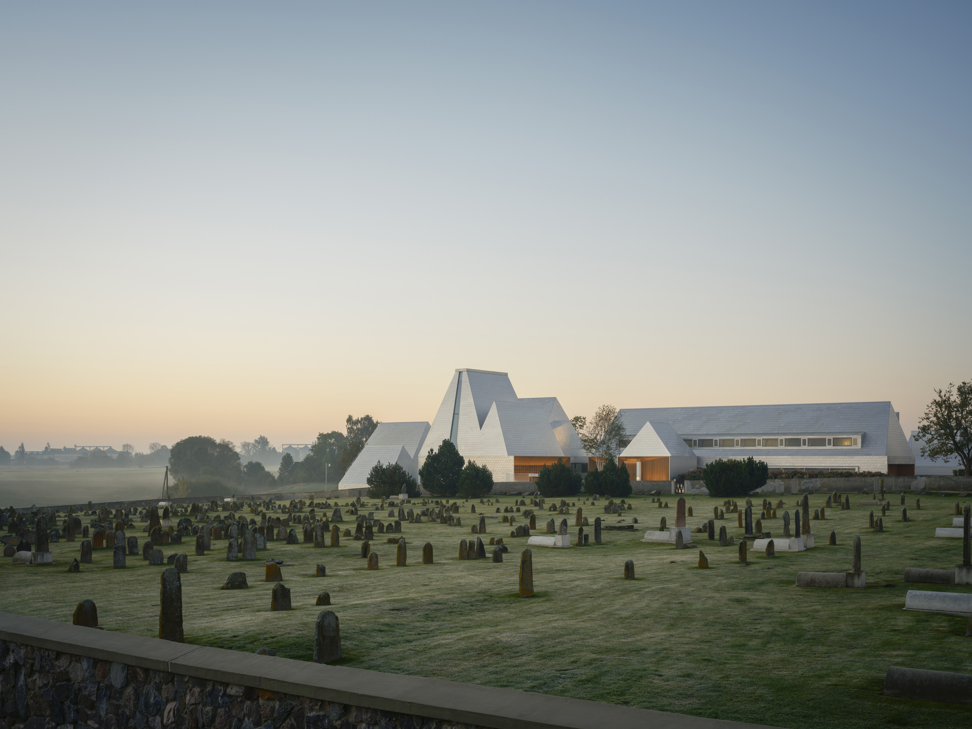 White faceted roof volumes rising above a cemetery at dawn with mist covering distant fields