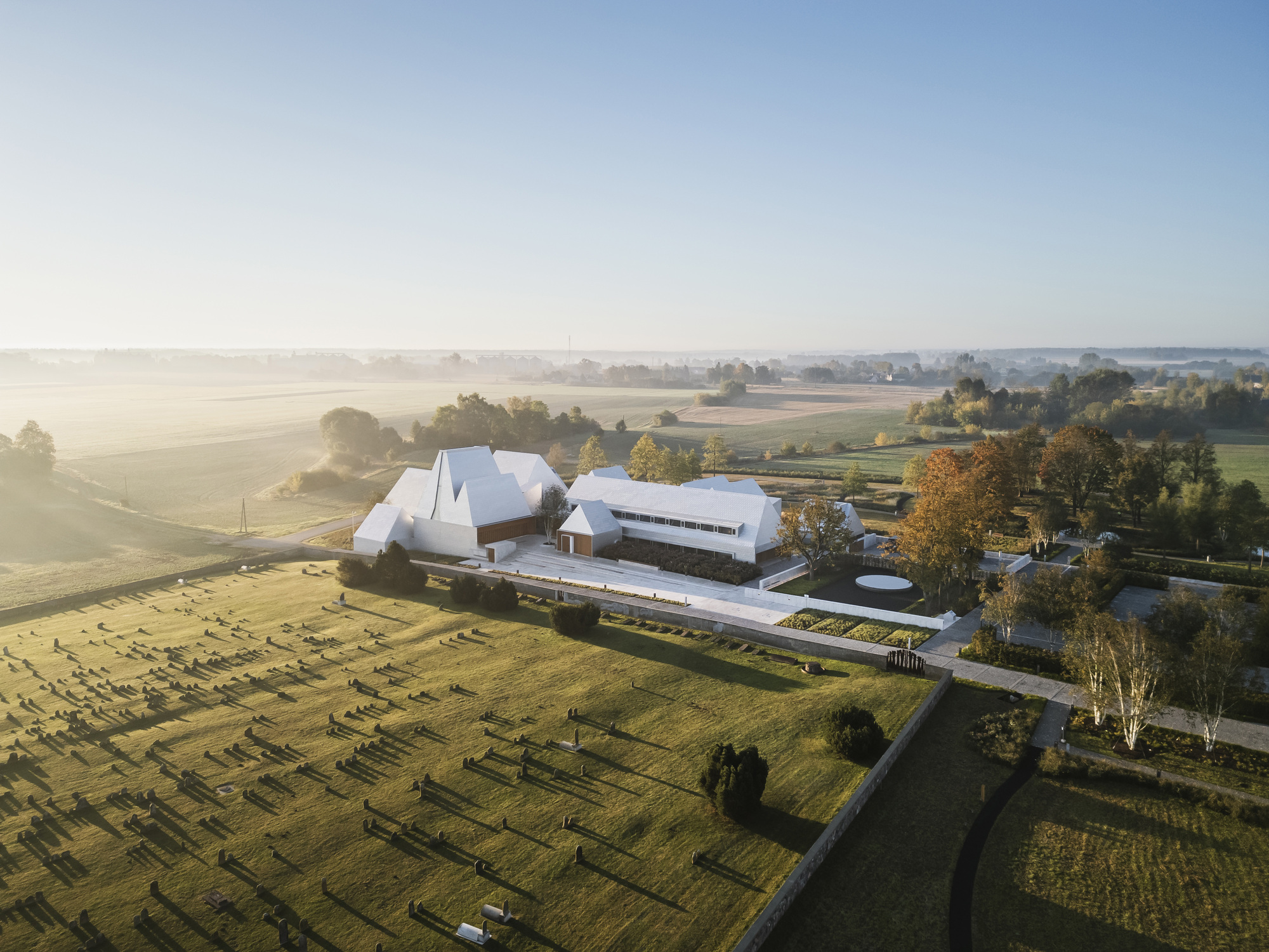 Aerial view of angular white roofs and linear wings bordered by a cemetery and morning fog