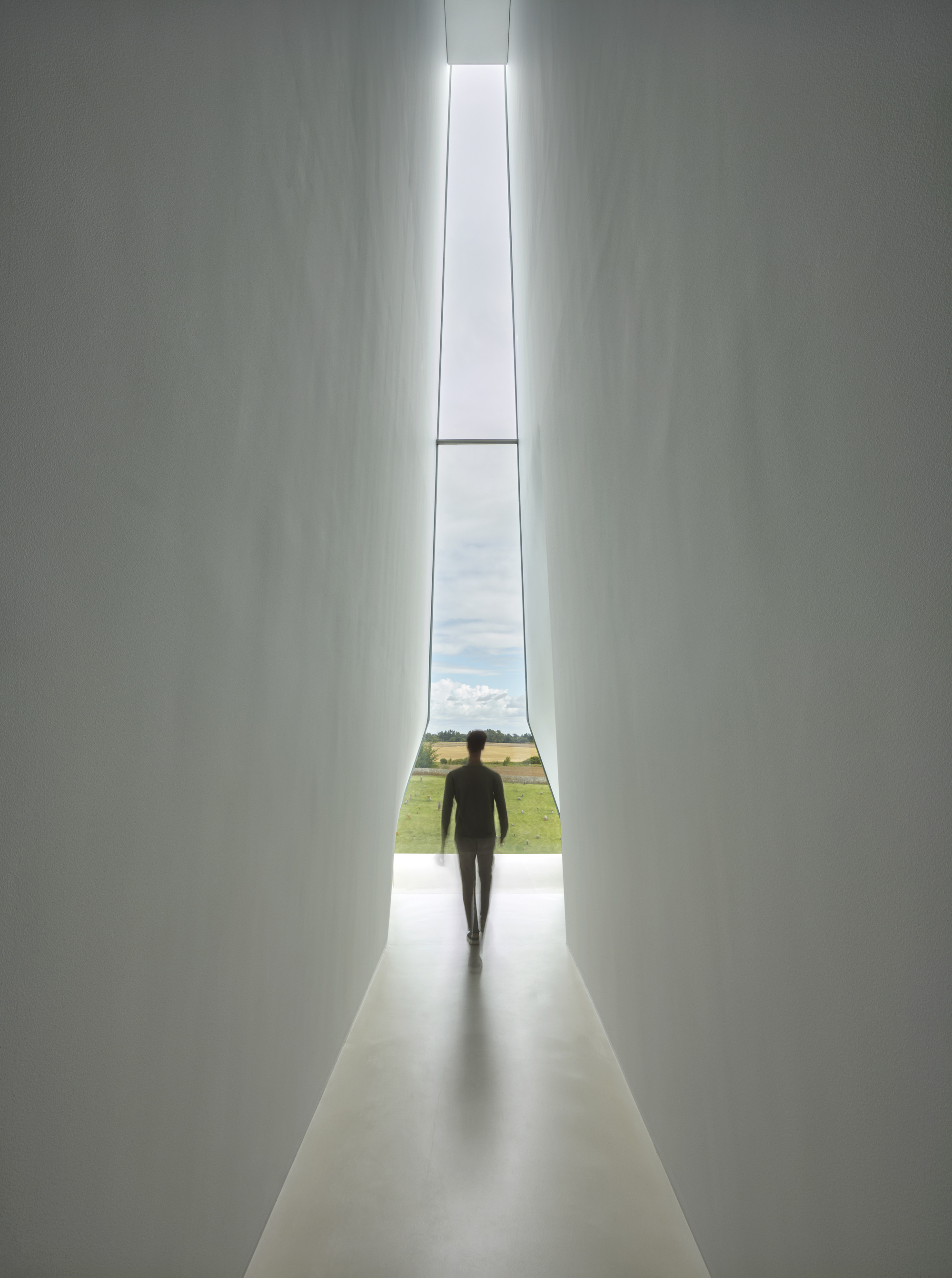 Tall vertical window slot framing a silhouetted figure walking toward countryside beyond white walls