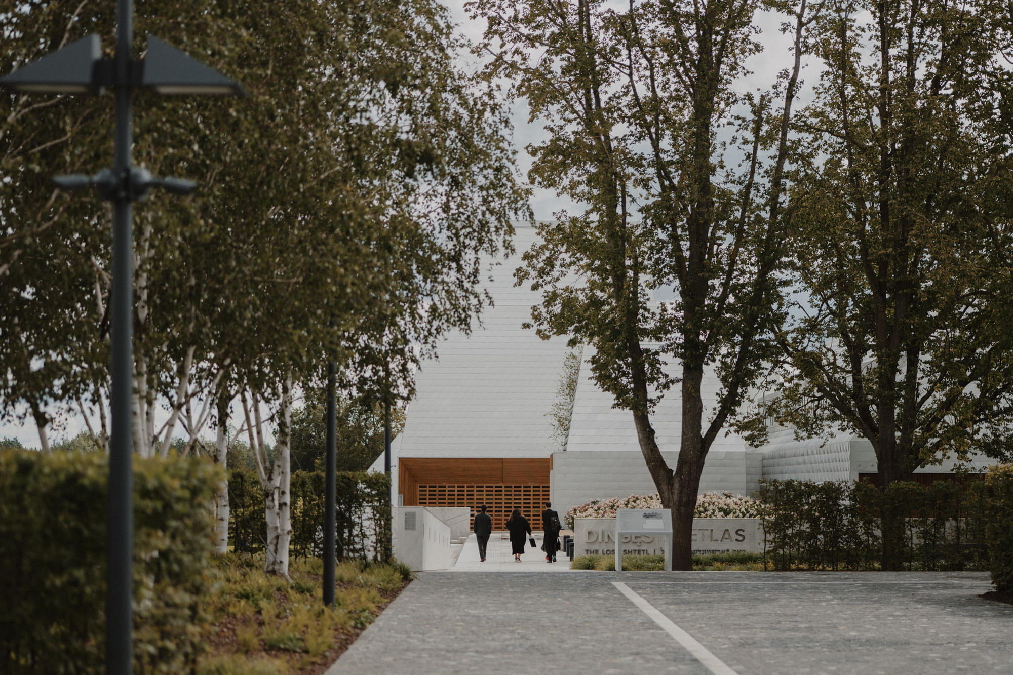 Entrance pathway with visitors approaching the white facade and timber canopy through trees