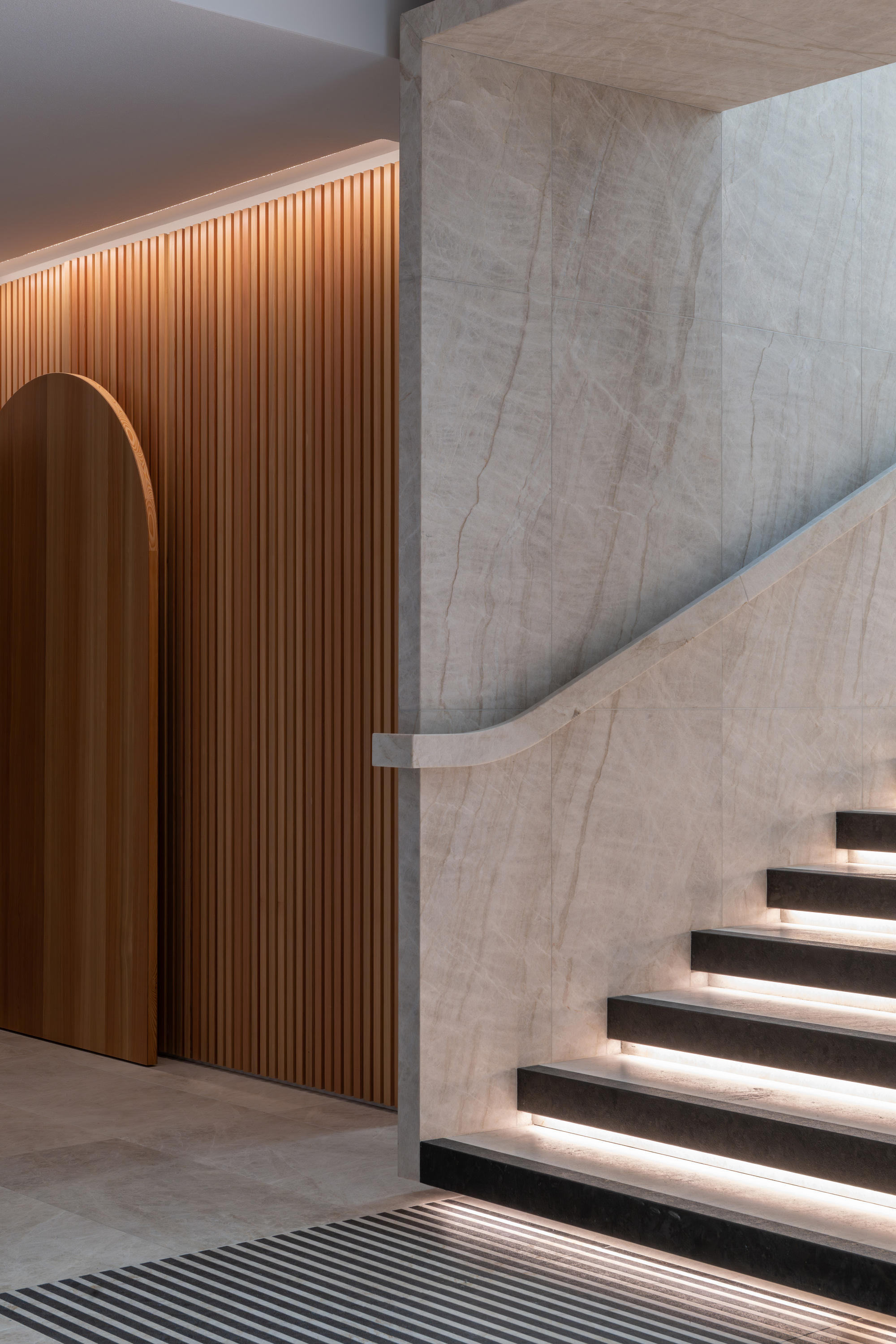 Junction of curved timber wall with arched opening and board-formed concrete stair with lit treads