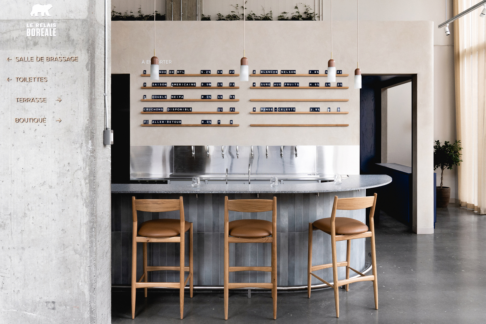 Curved bar with fluted tile base, oak stools, and wall-mounted shelving beside concrete directional signage panel