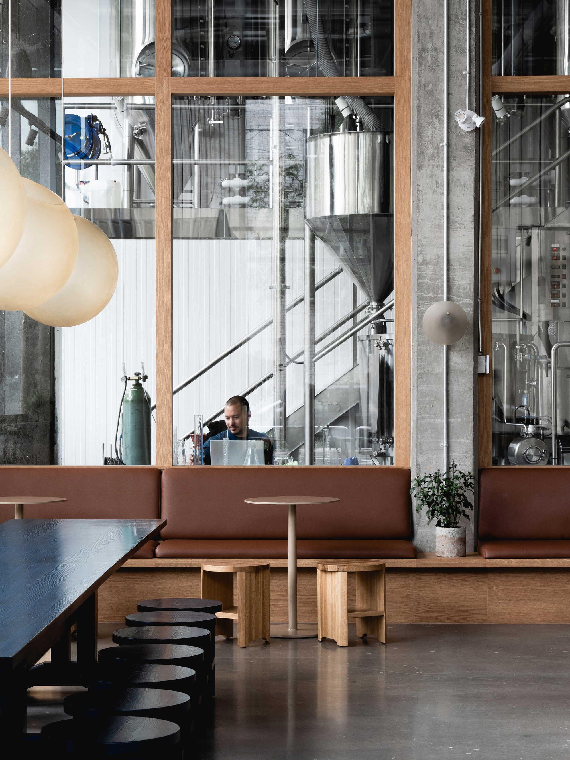 Timber-framed glazed wall reveals brewing equipment and a figure on the staircase beyond the banquette seating