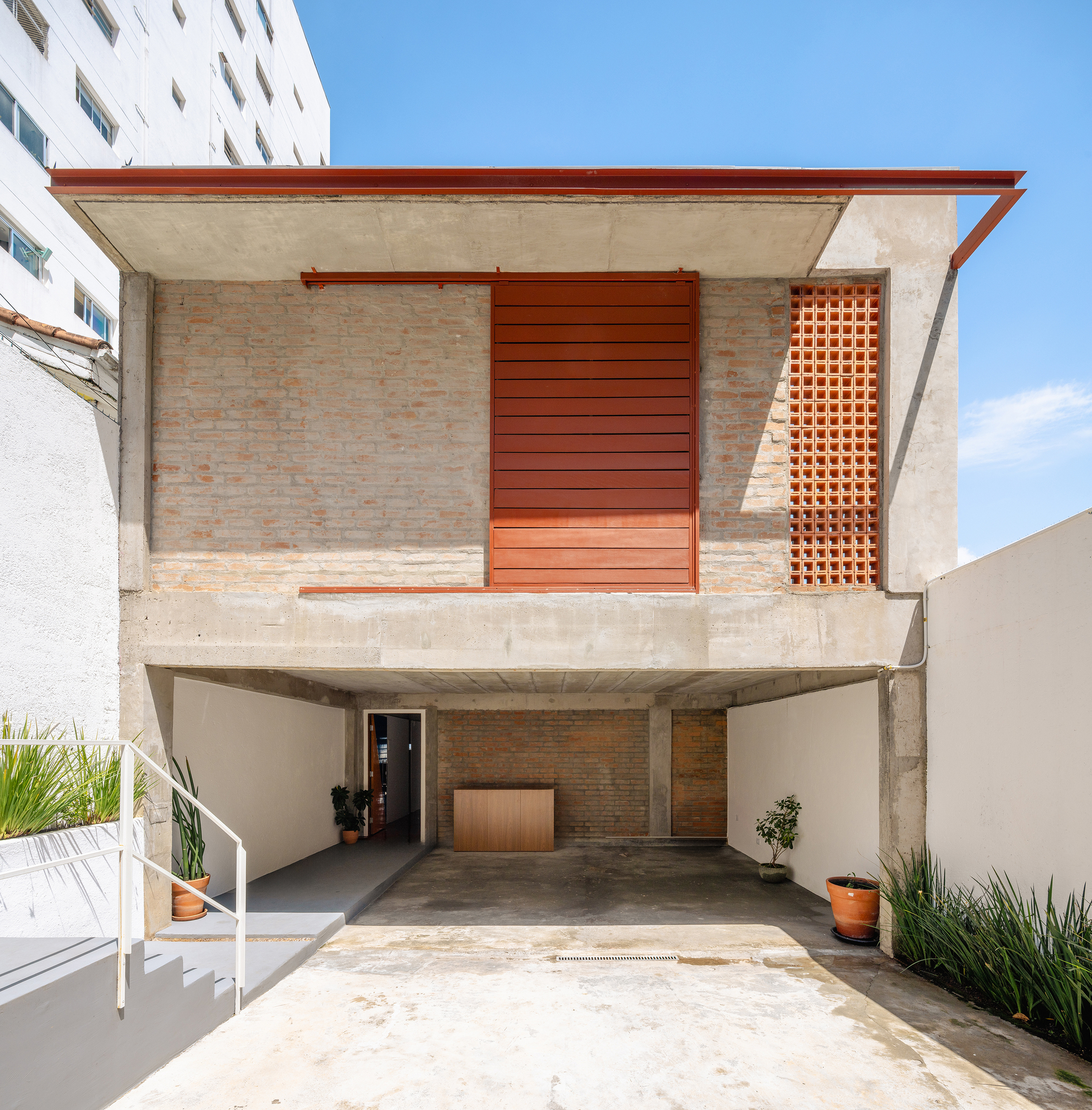 Two-story facade with exposed concrete, brick walls, sliding orange timber panels and carport below under clear sky