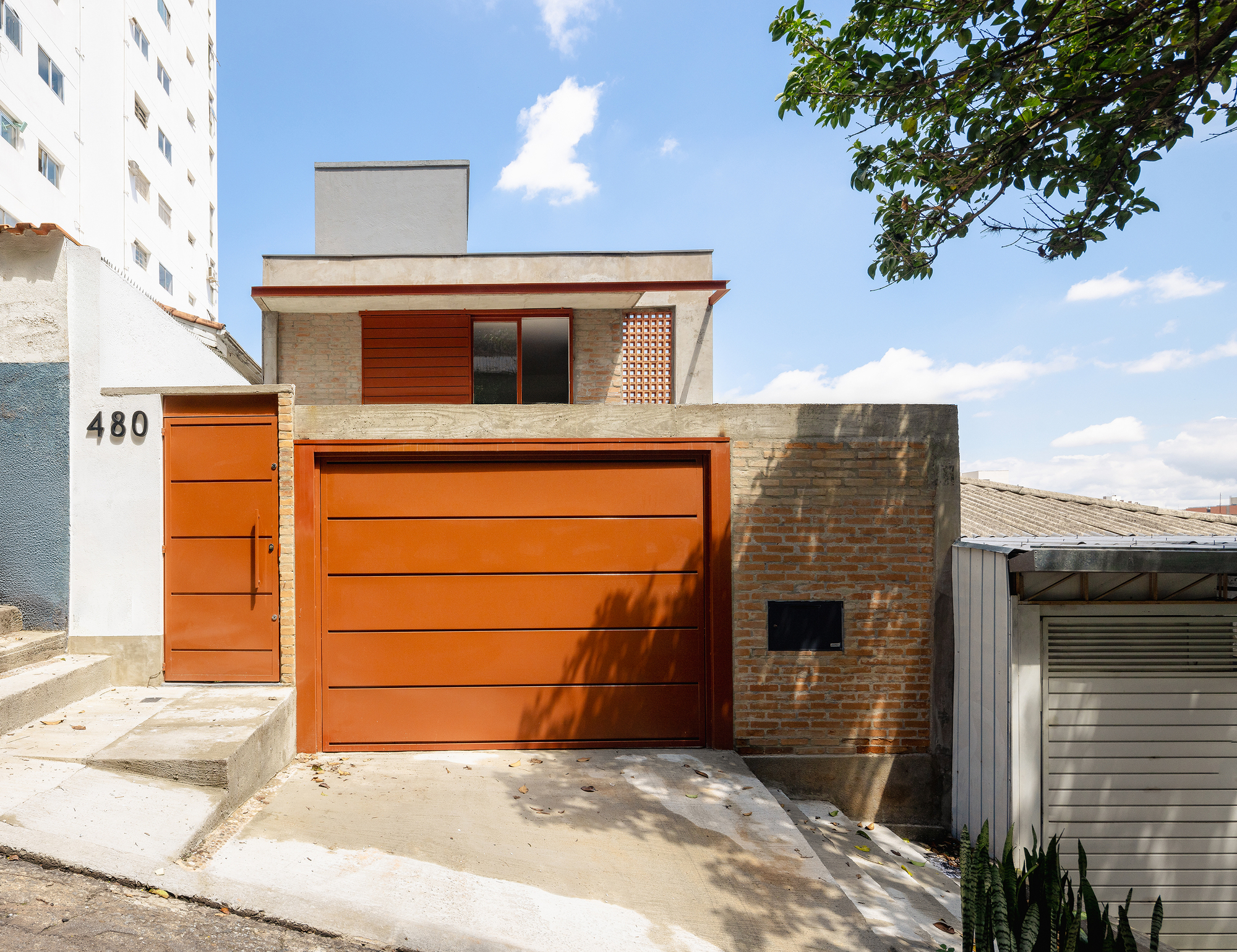 Street view showing orange timber garage door, brick volumes and lattice screen with tree shadow on pavement