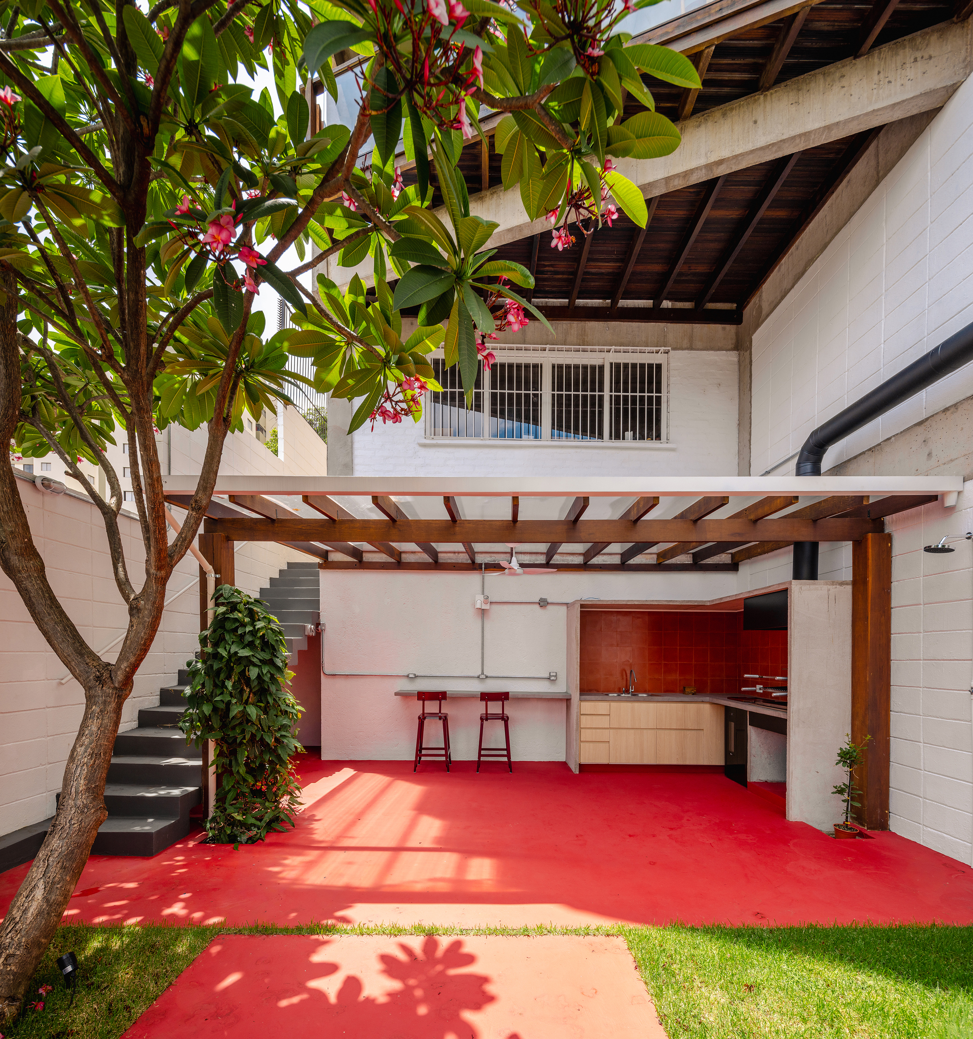 Courtyard with red resin floor, flowering frangipani tree, and timber pergola shading an outdoor kitchen