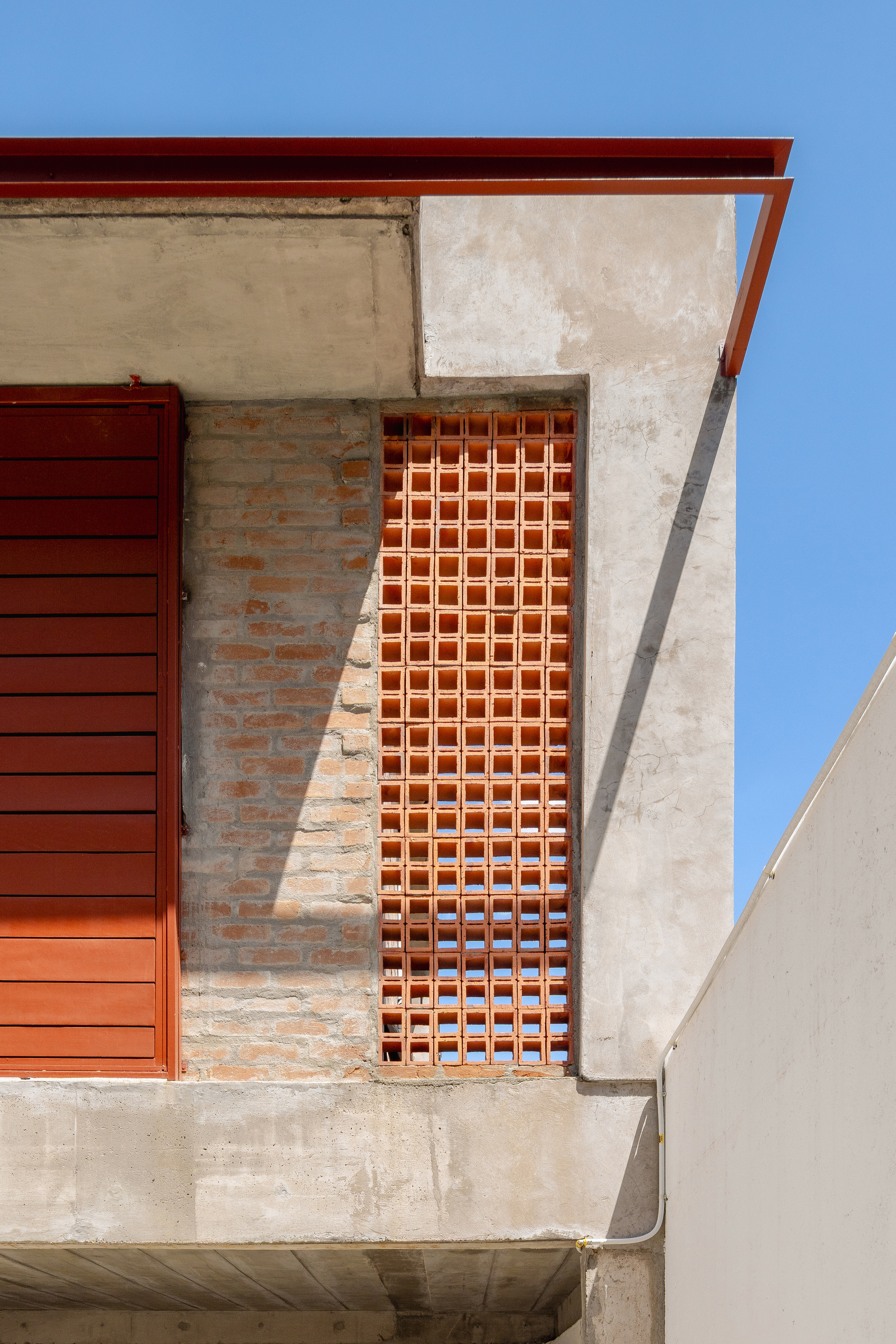 Terracotta brick screen window set into exposed concrete and weathered plaster facade under clear blue sky