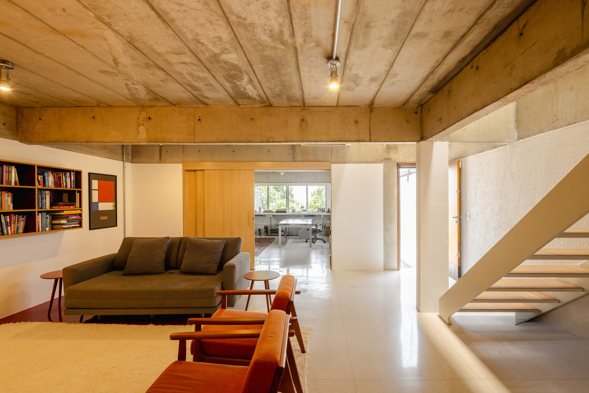 View through the living room toward a daylit workspace with concrete staircase on the right