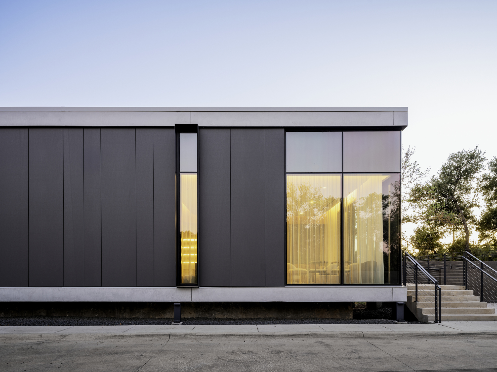 Dark metal panel facade with lit vertical slot window and larger glazed opening at dusk