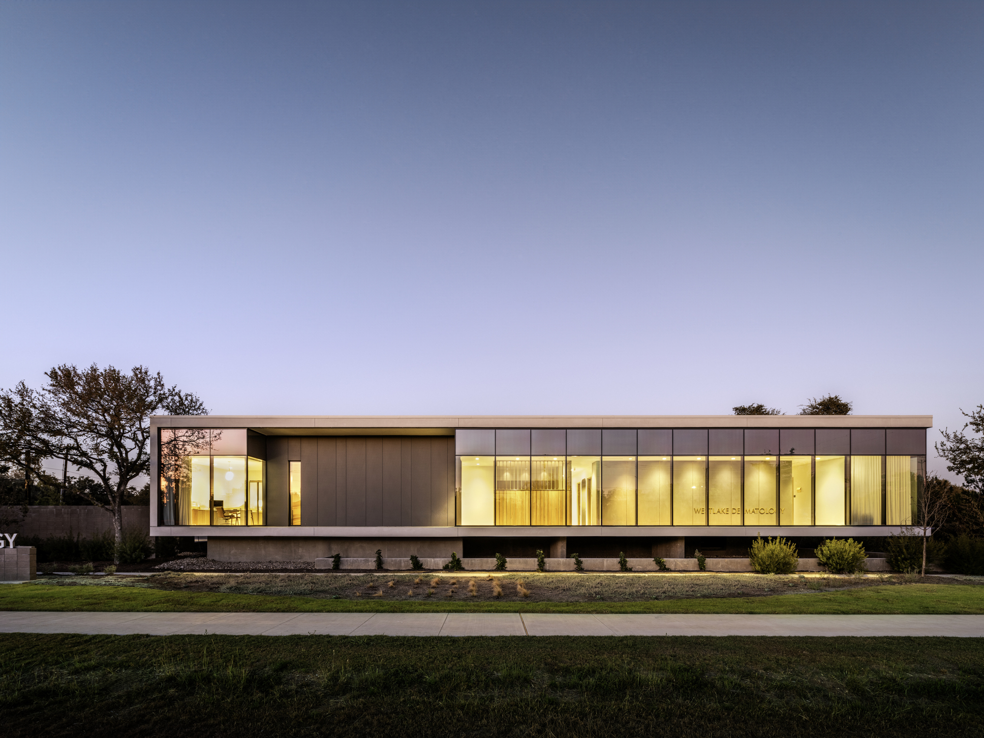 Long view of the glass and metal facade glowing at twilight behind lawn and trees