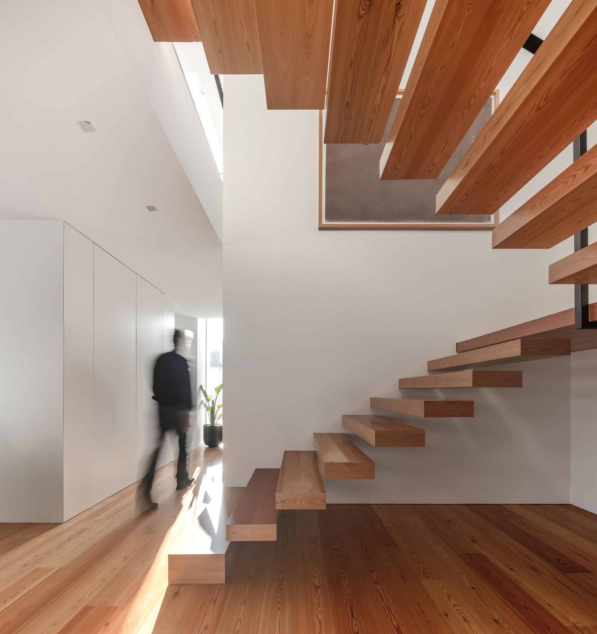 Cantilevered timber staircase with floating treads alongside built-in white cabinetry and a blurred figure below