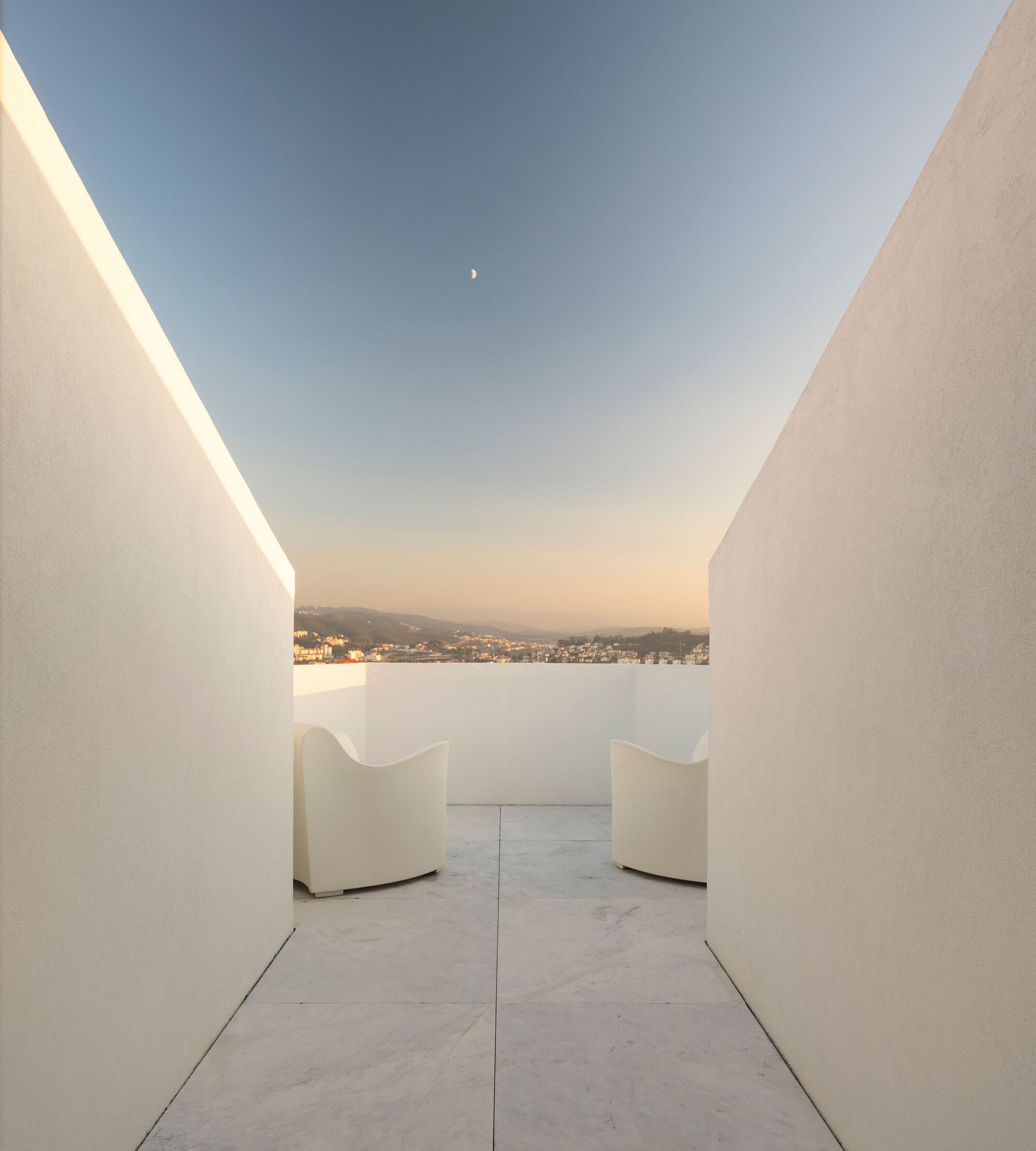 White rendered passage with two curved chairs framing a view toward distant hills at dusk