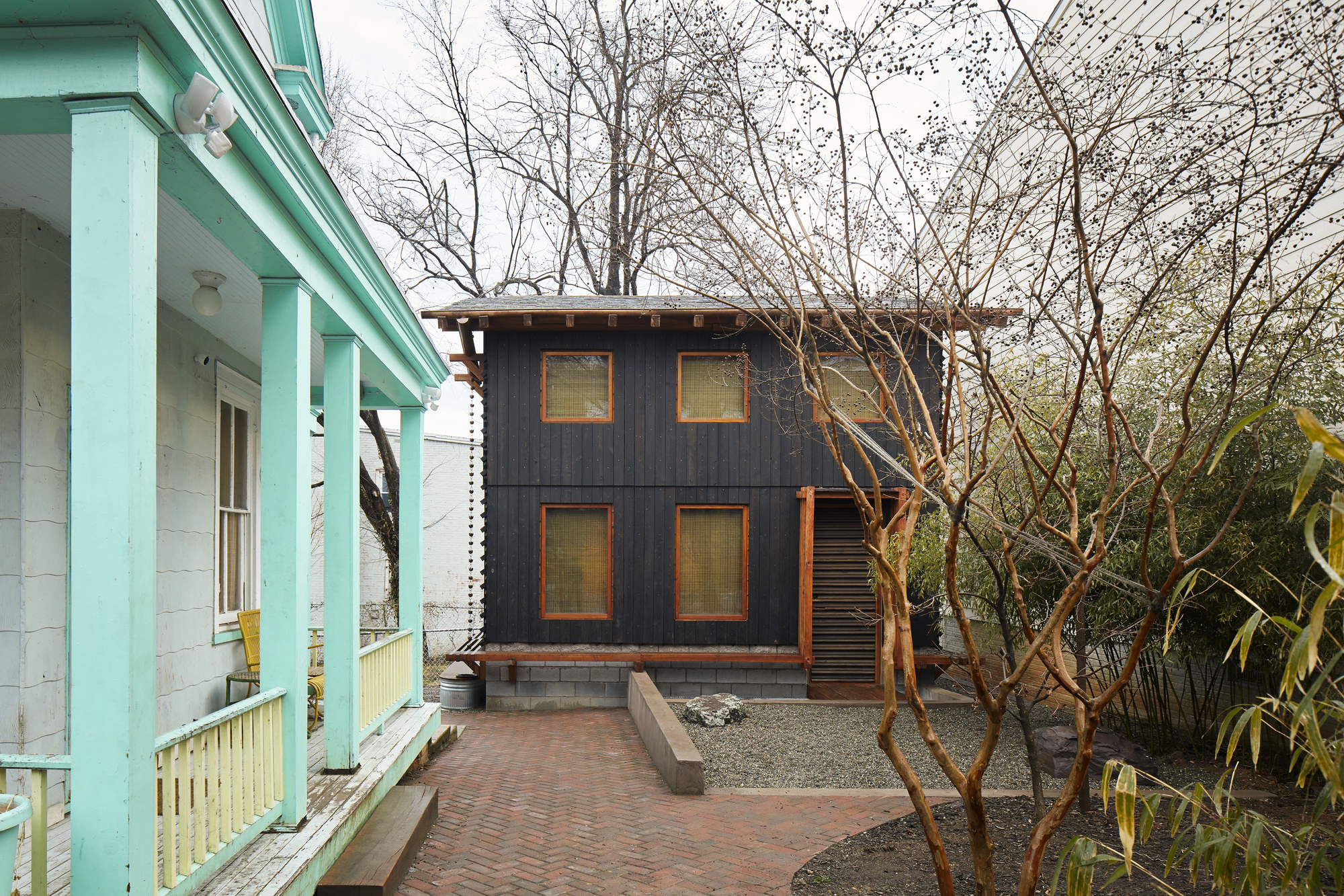 Two-story dark timber-clad volume with square windows seen from a turquoise porch across bare trees