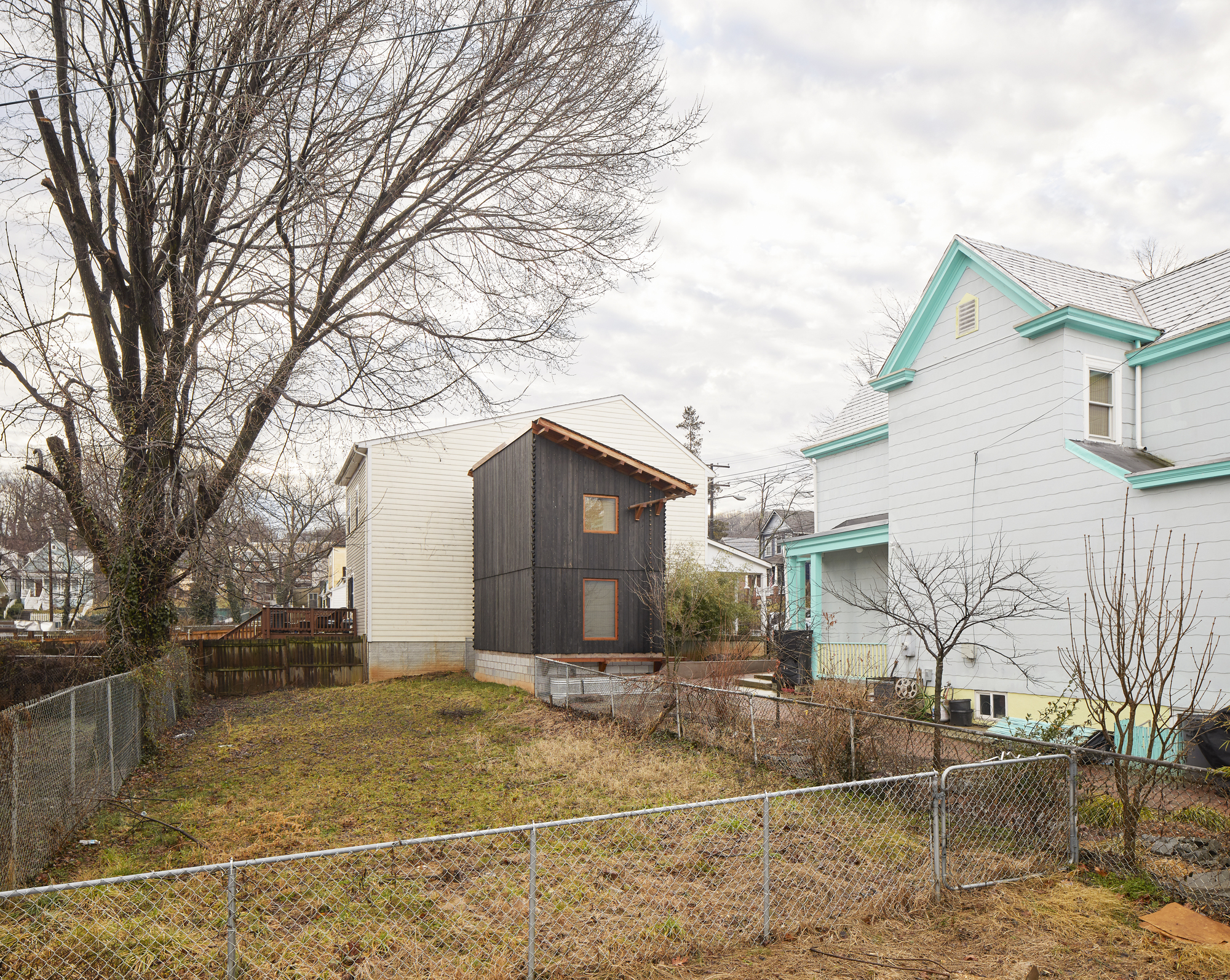 Side view of the compact timber volume on a sloped lot between neighboring clapboard houses