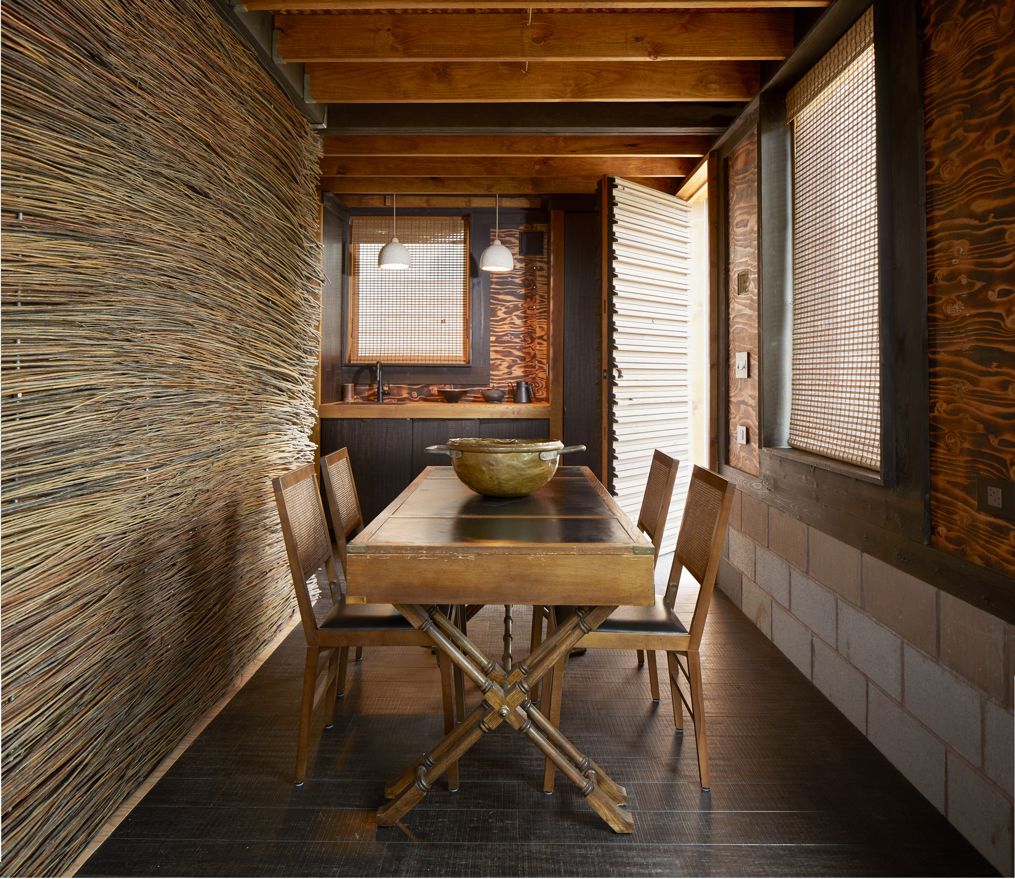 Dining area with exposed timber beams and layered wall treatments of woven screens and plywood panels