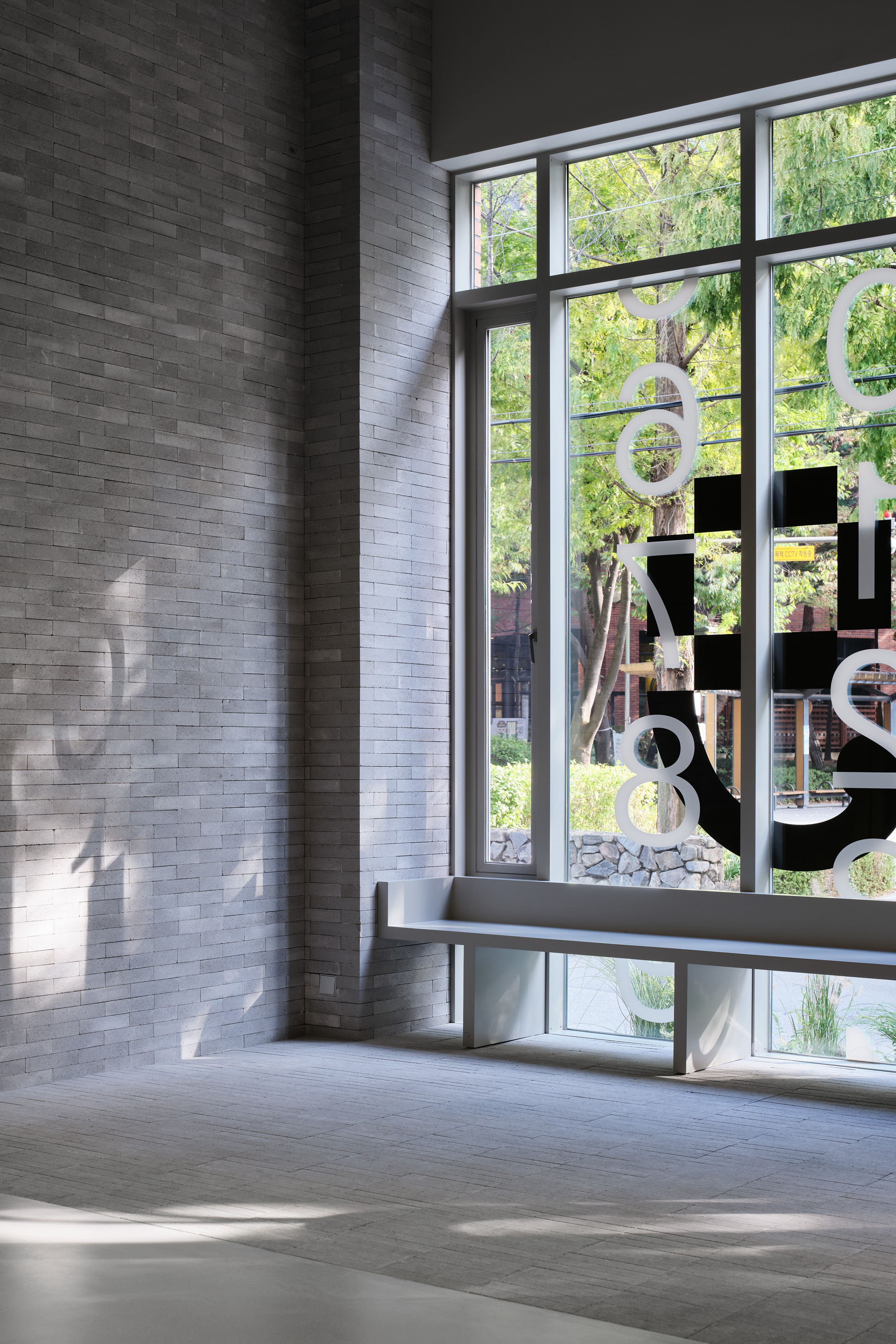 Grey brick wall beside a full-height window with graphic signage overlooking a sunlit courtyard