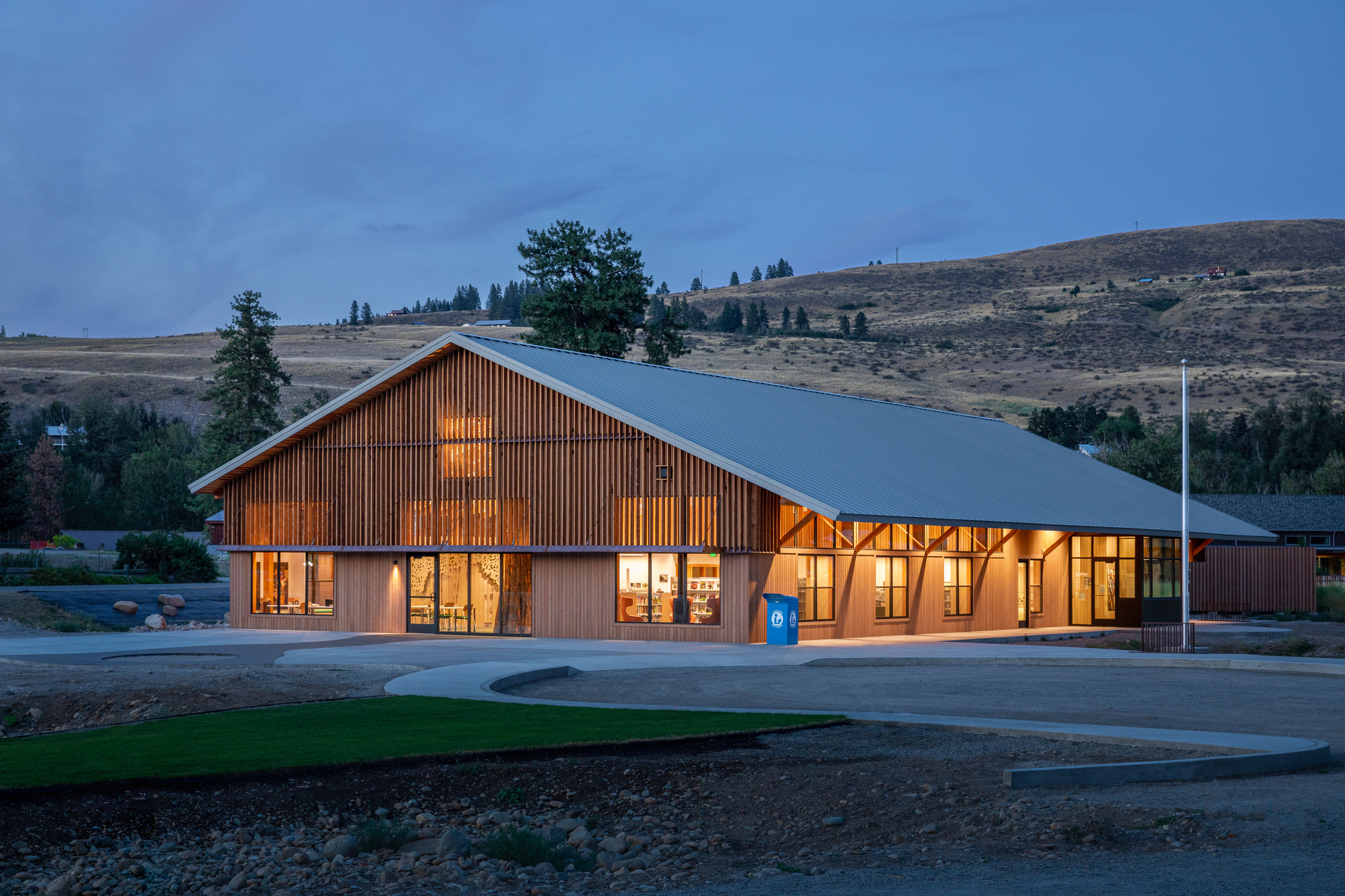 Gabled timber-clad library building with illuminated glazed facade at dusk in a rural hillside setting