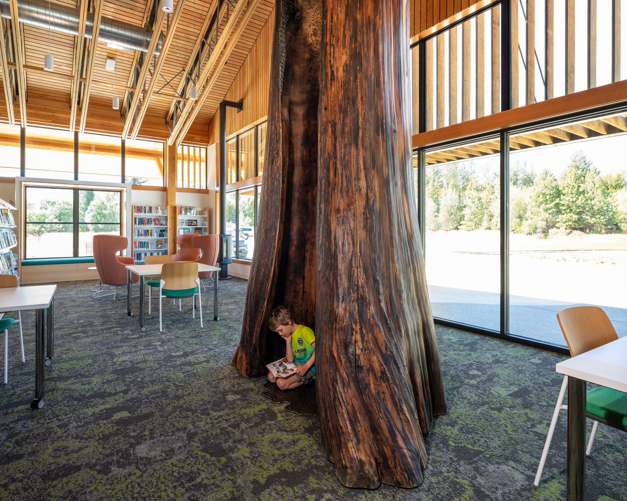 Interior library space with exposed timber trusses and a large preserved tree trunk with a child reading at its base