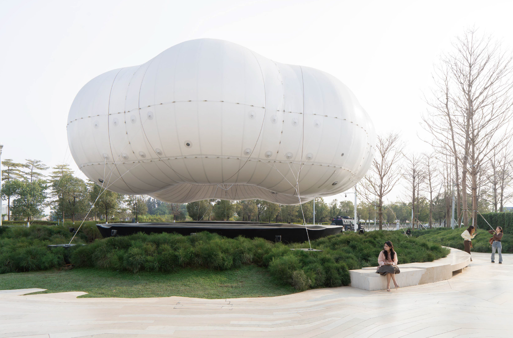 White inflatable balloon structure tethered above planted berms with visitors seated on concrete benches below