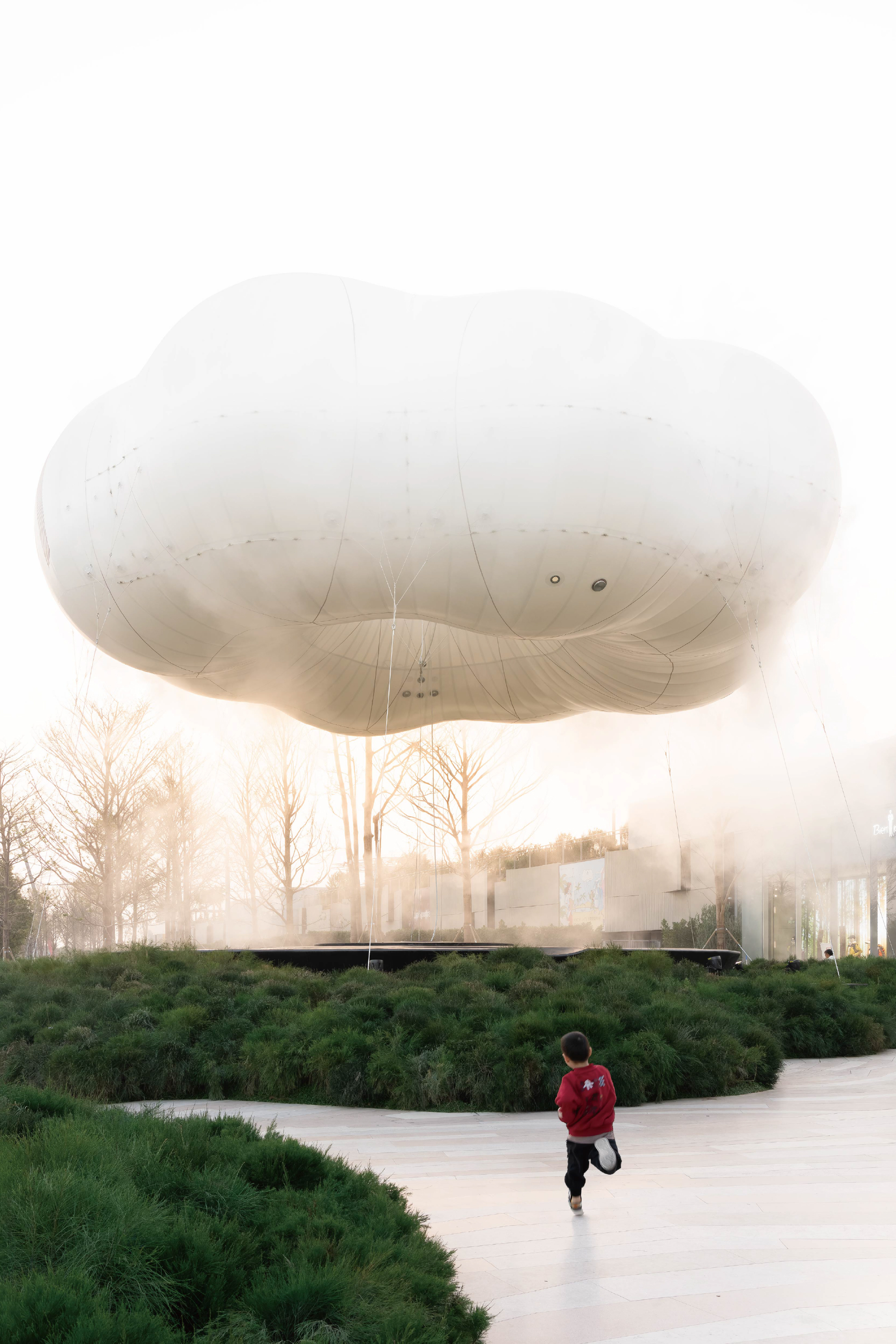Tethered white balloon hovering in fog with a person in red jacket walking on paving below
