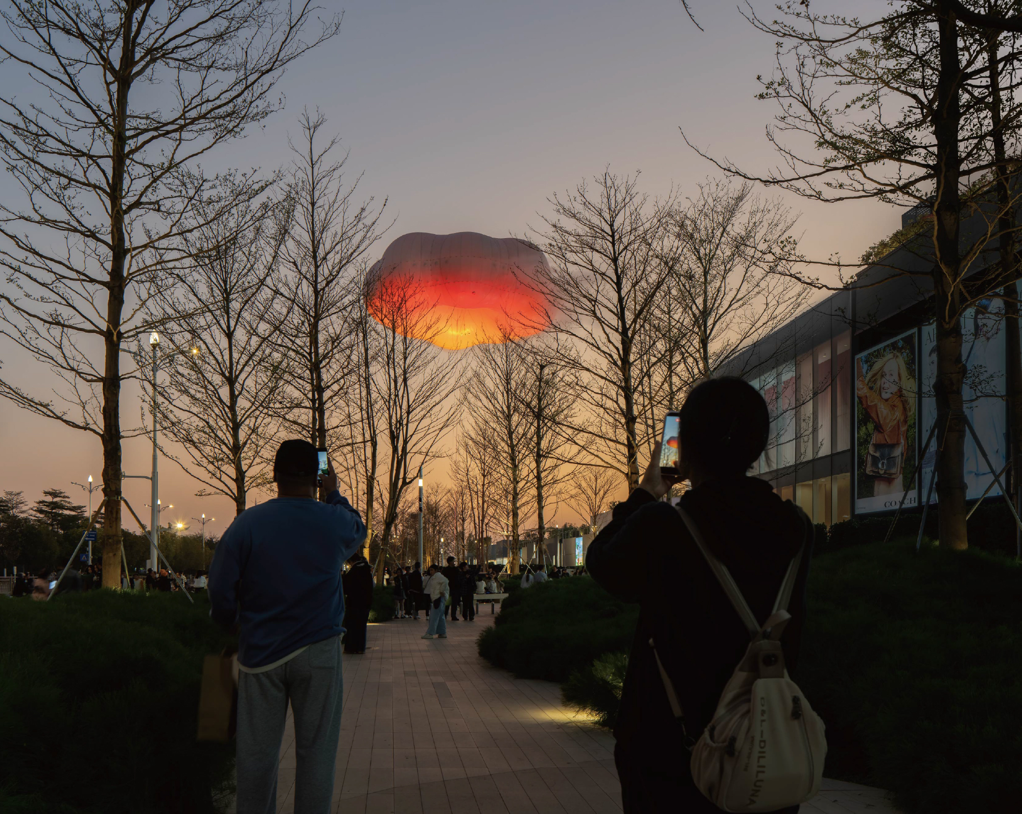 Illuminated red and orange balloon glowing at dusk above bare trees with silhouetted figures photographing below