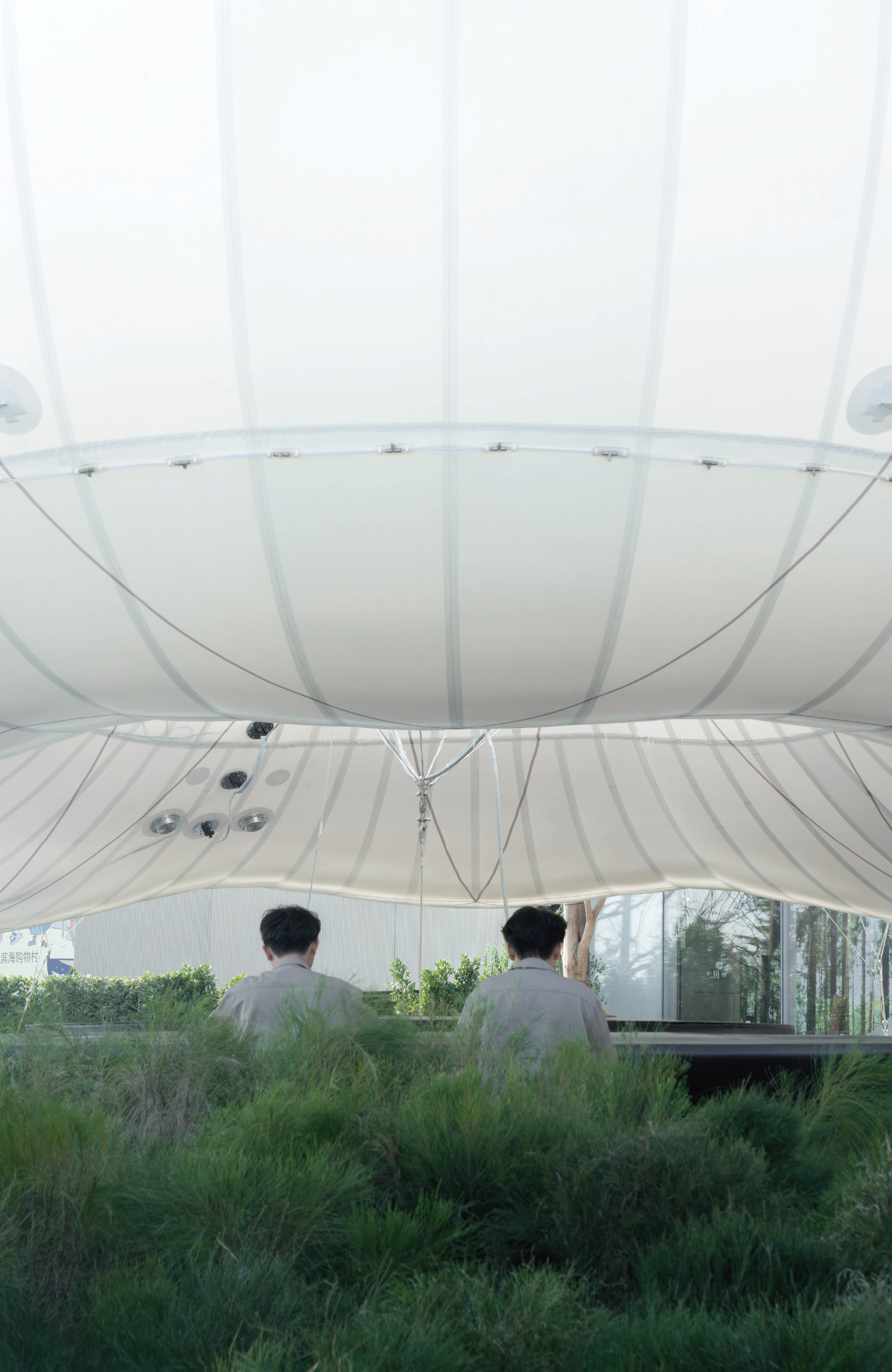 View from inside white tensioned fabric envelope looking down at two figures seated in planted grasses