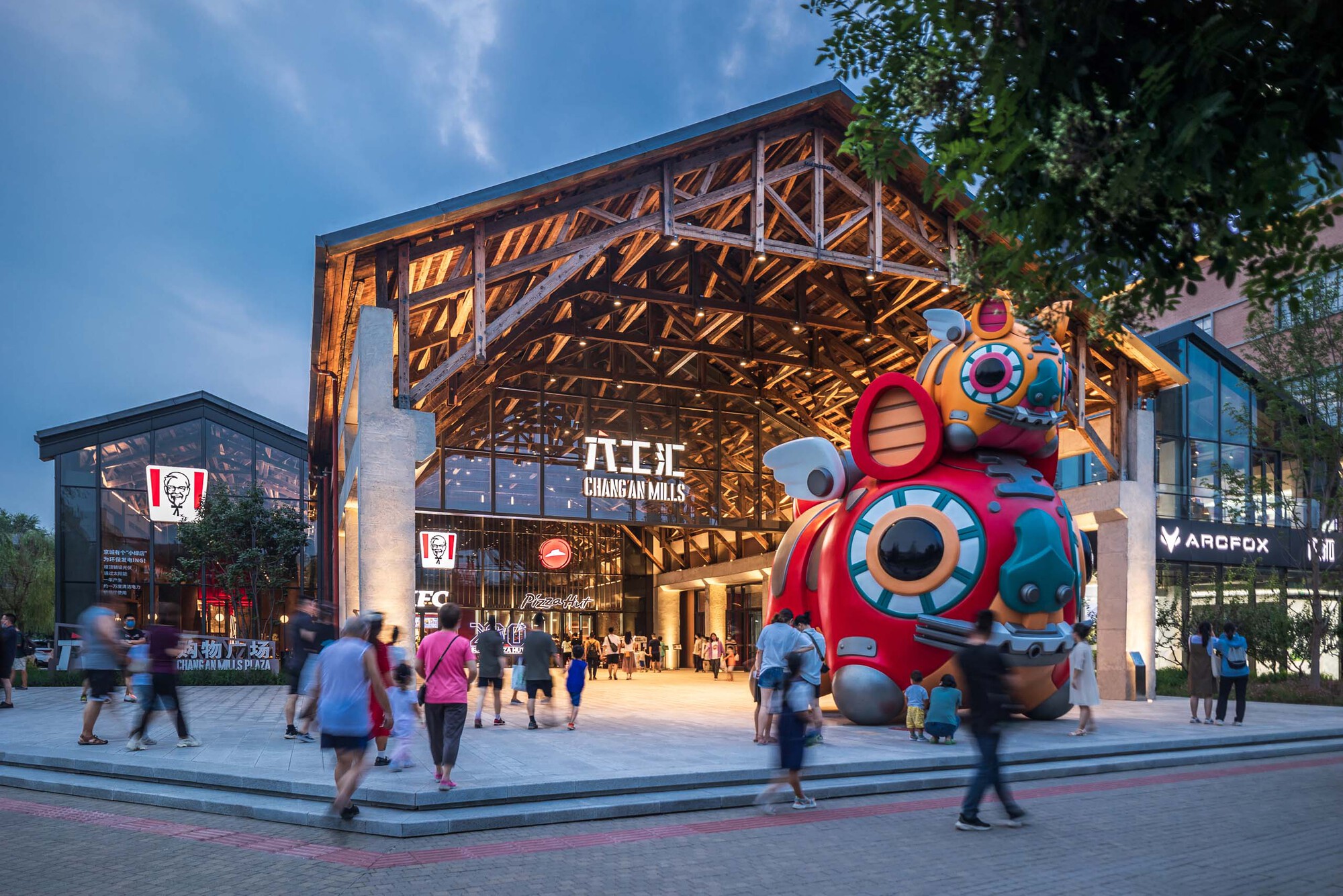 Exposed timber truss canopy over a plaza entrance with pedestrians and a colorful sculpture at dusk