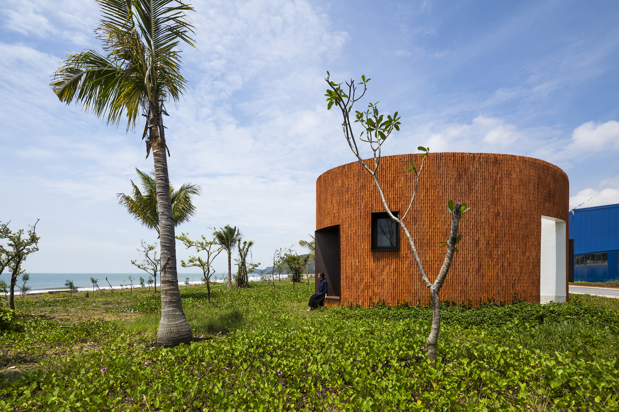 Curved brick volume with small window and door openings amid palm trees and coastal vegetation
