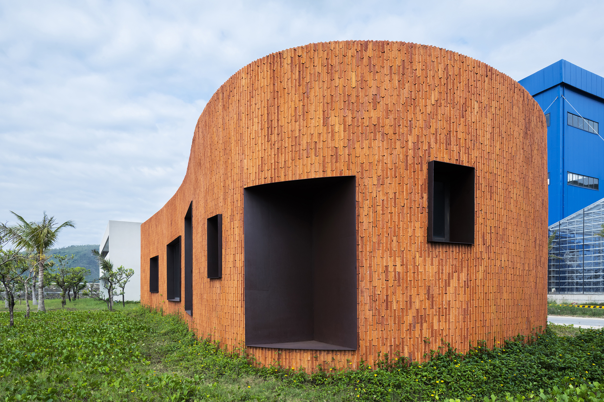 Curved brick facade with deep-set rectangular openings in a grassy field under cloudy skies