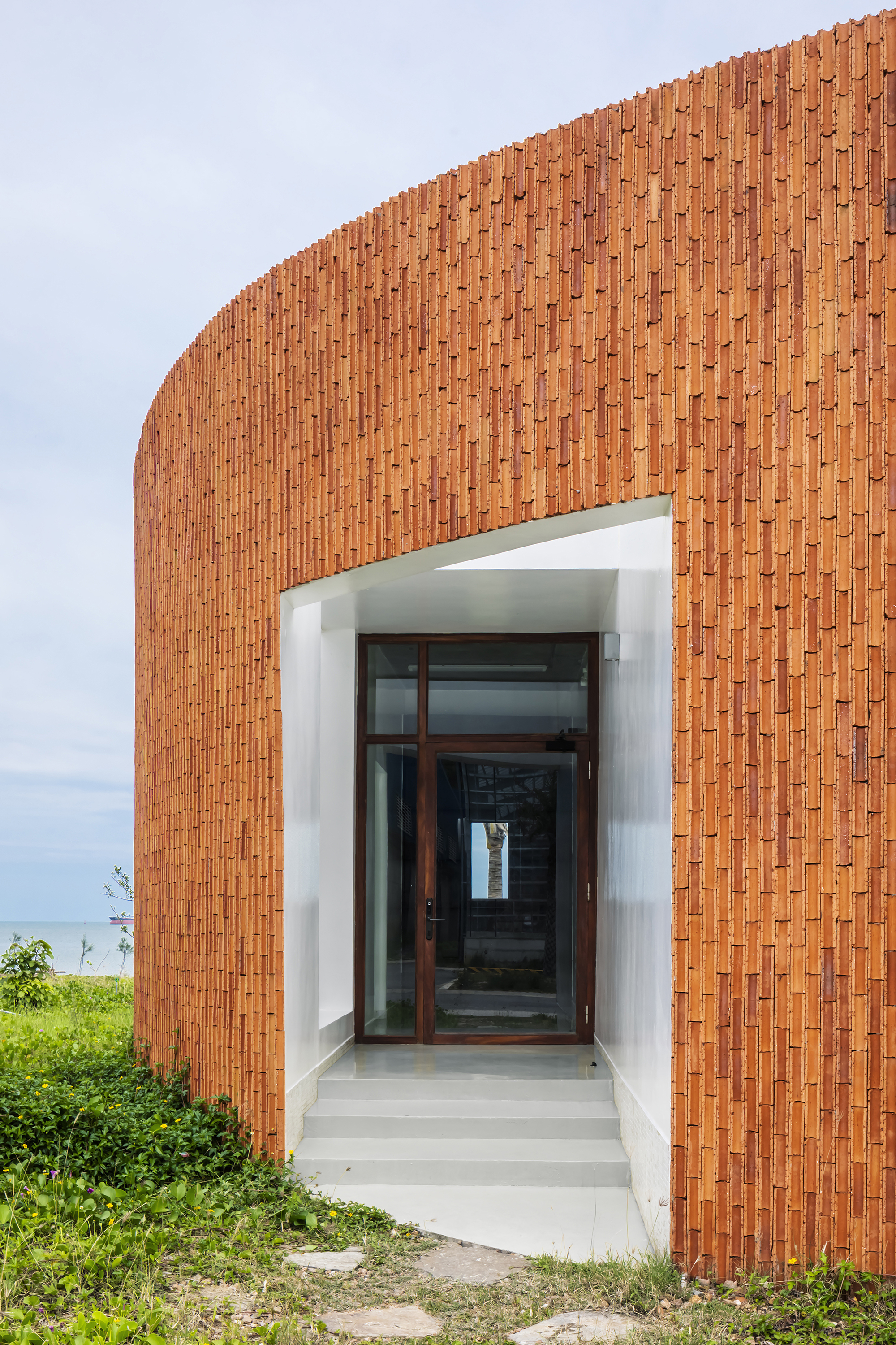 Recessed entrance with white concrete steps through curved terracotta brick wall facing coastal vegetation
