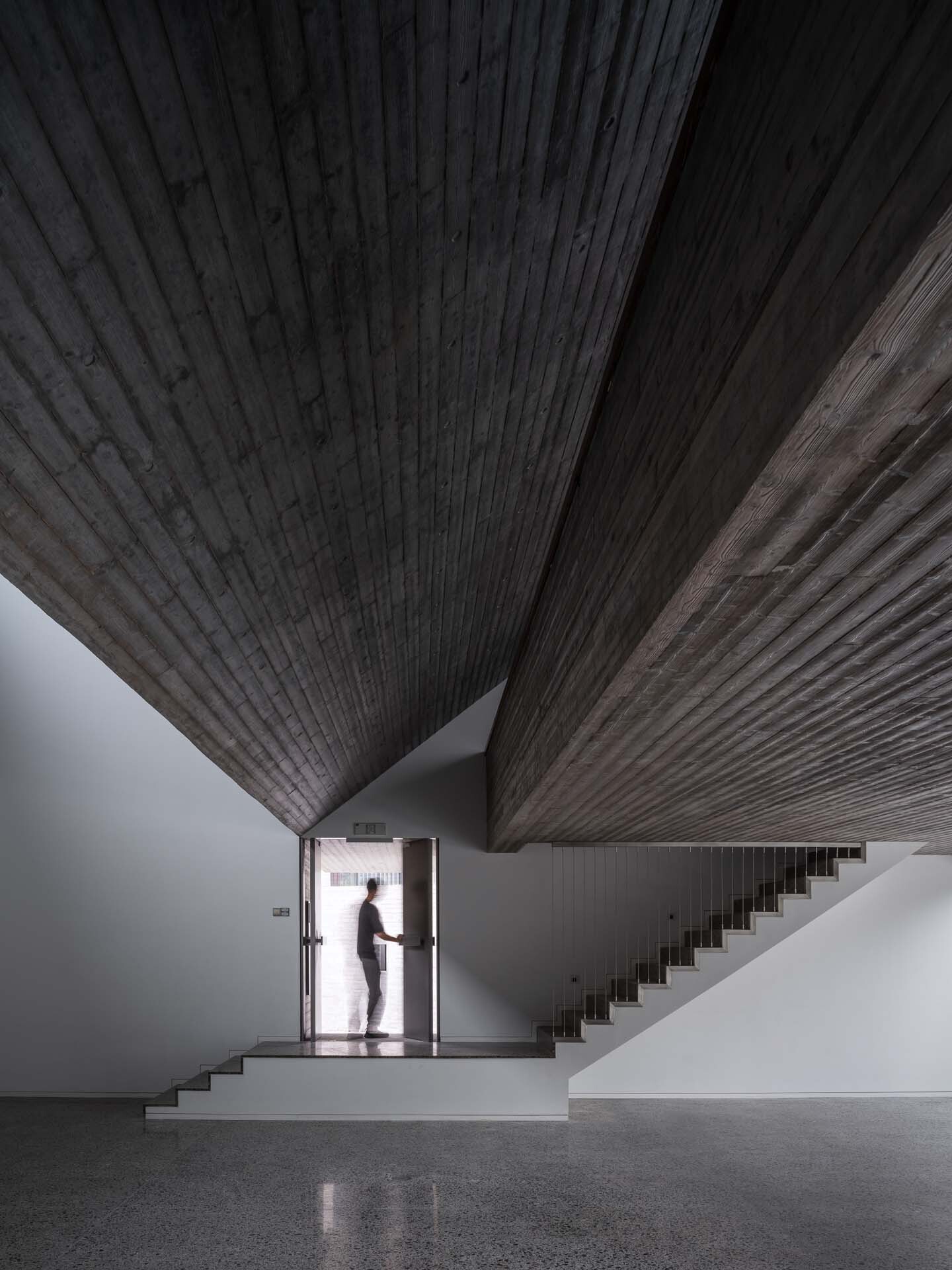 Double-height entry hall with angular timber ceiling planes and a figure silhouetted in the doorway