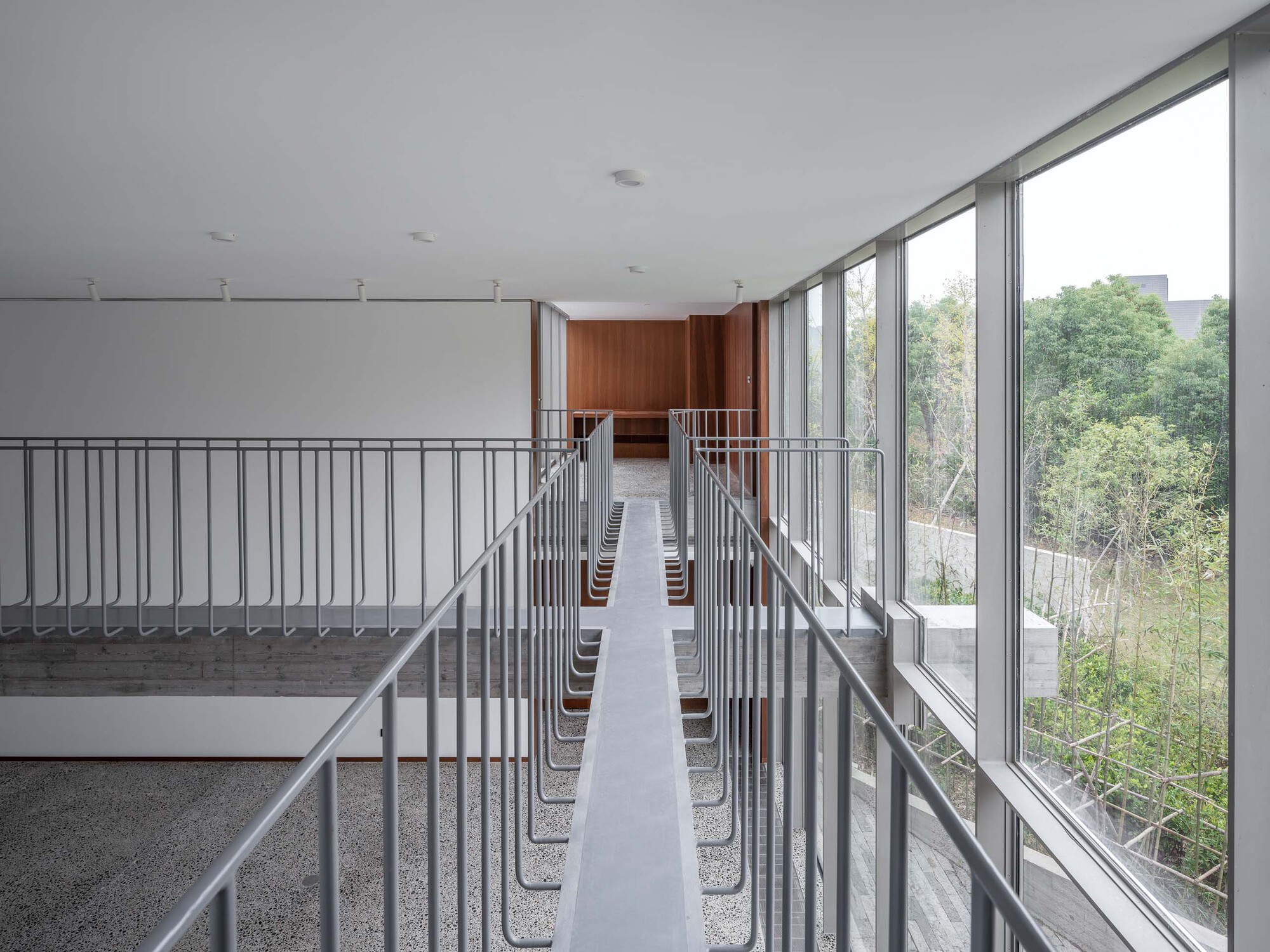 Narrow interior bridge with metal handrails and rows of seating leading toward timber-clad volume