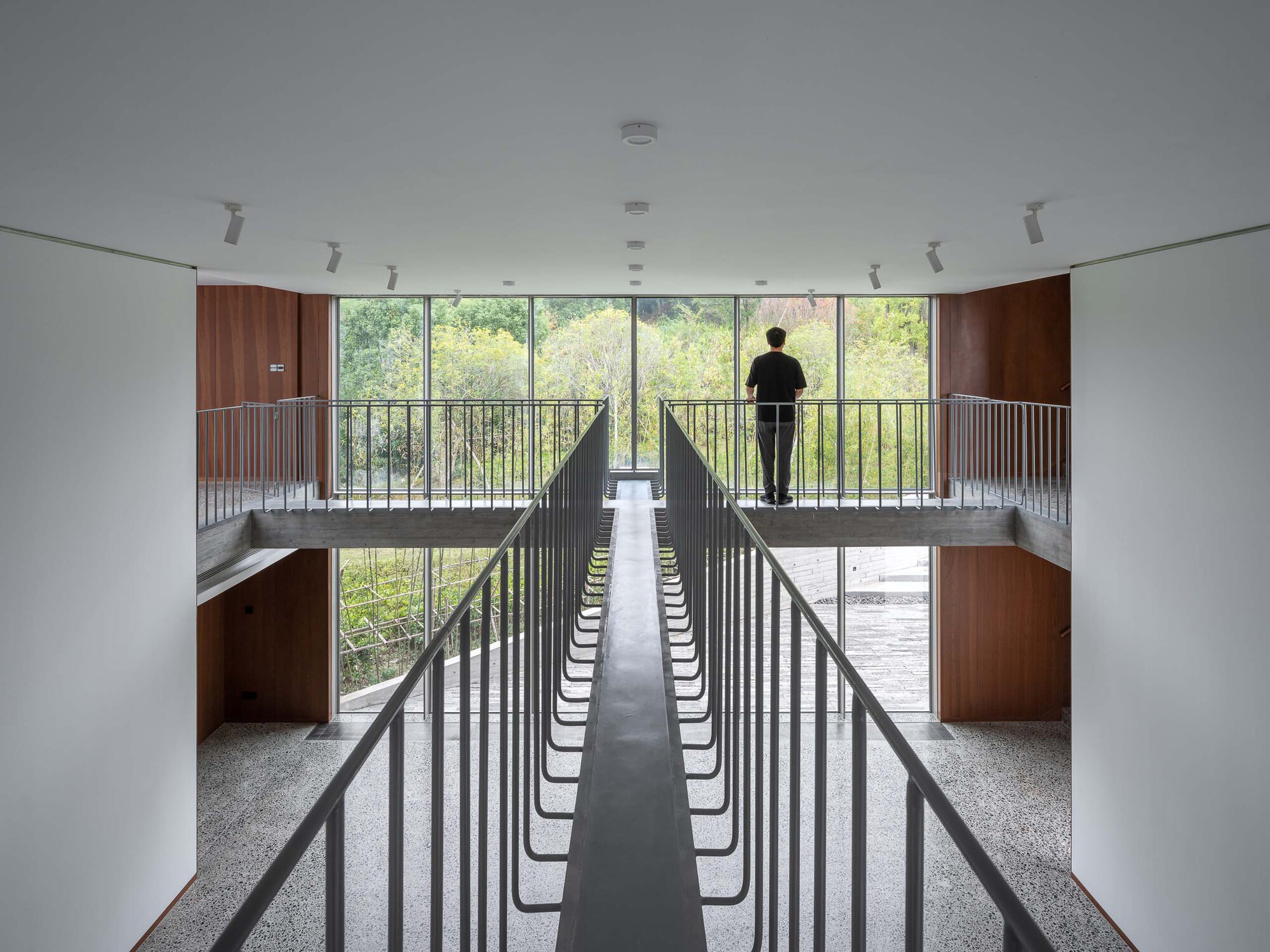 Double-height interior with vertical black railings and a figure silhouetted at the glazed balcony overlooking trees