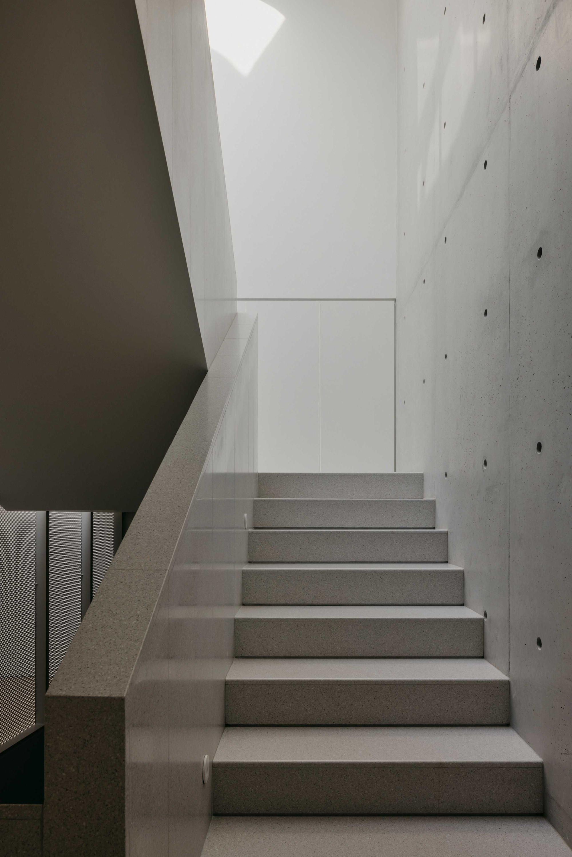 Concrete staircase rising between exposed concrete walls with circular formwork ties under a skylight