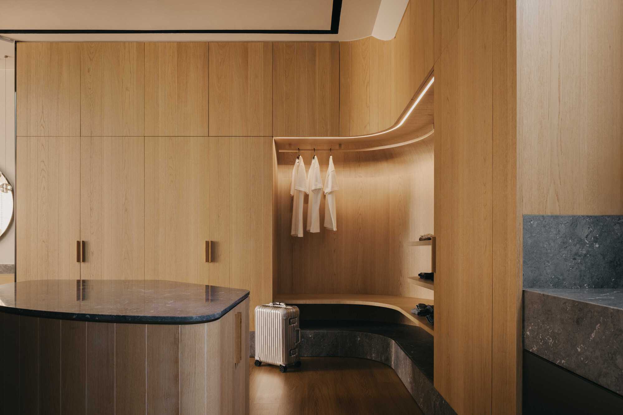 Wardrobe with curved timber hanging rail, integrated lighting, and stone bench seating beside a suitcase