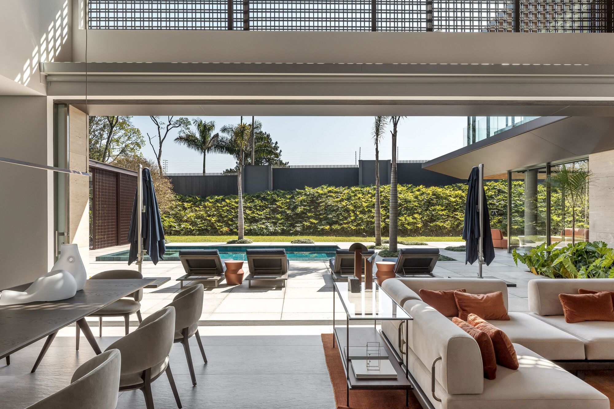 Double-height living room opening to pool terrace with palm trees and hedge wall beyond