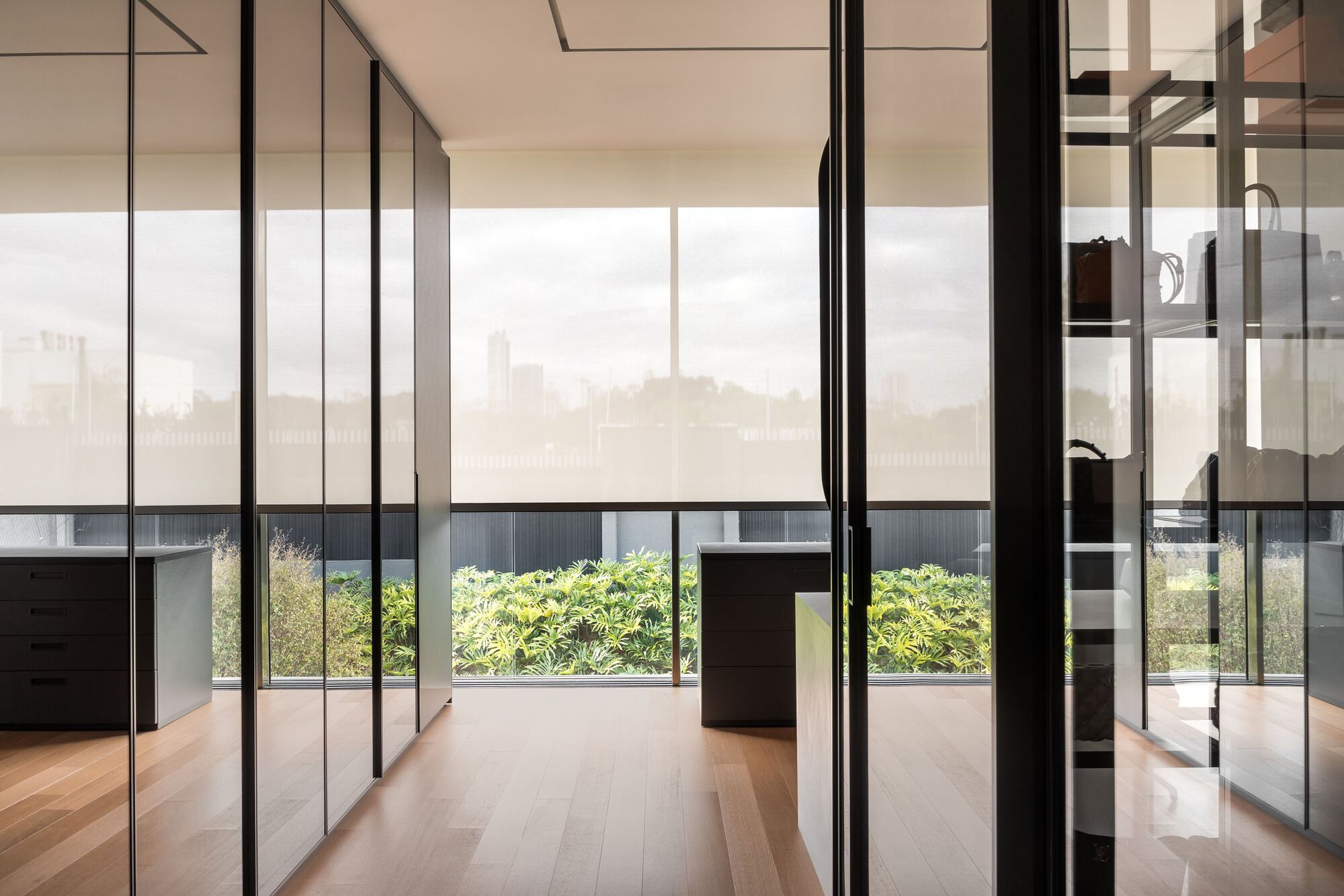 Glass-partitioned hallway with wooden flooring overlooking a terrace planted with low shrubs under hazy sky