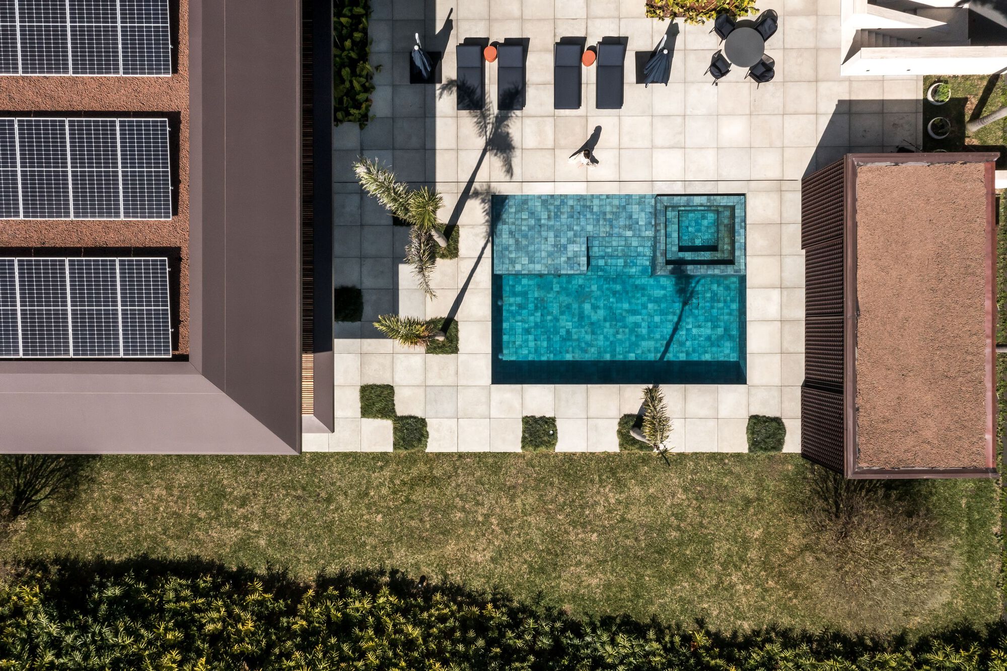 Aerial view of the tiled pool deck with lounge furniture surrounded by white pavers and landscaped beds