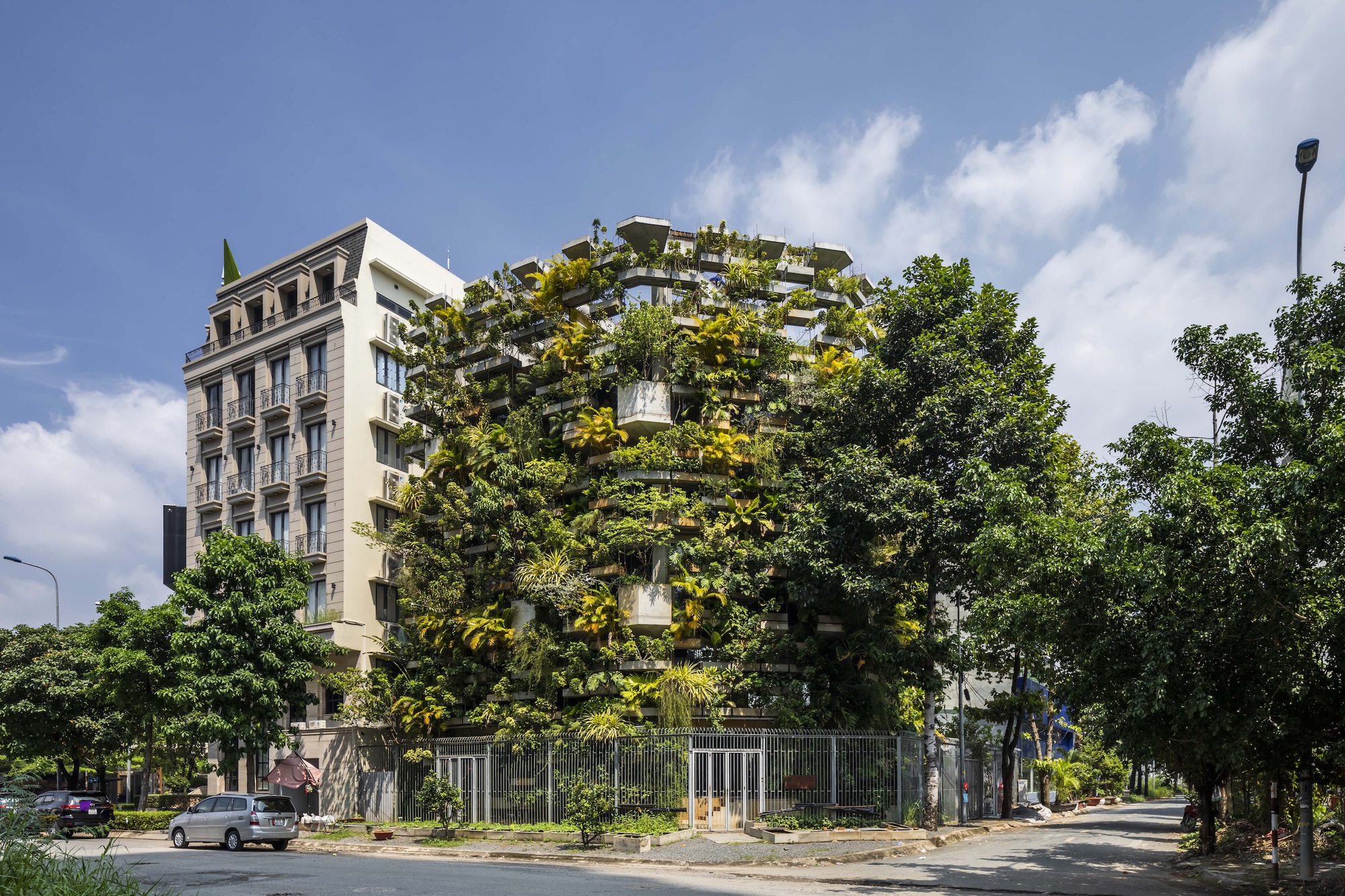 Street corner view of the planted facade tower rising behind tree-lined sidewalks under afternoon clouds