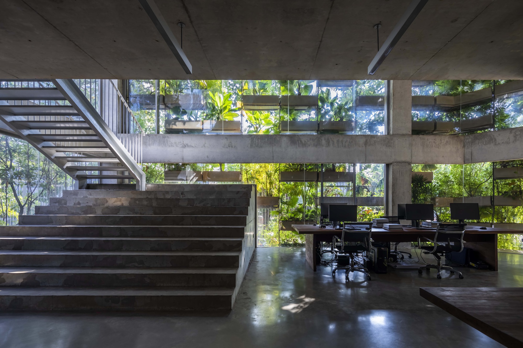 Concrete staircase rising beside open workspace with desks below planted balcony edges visible through glazing