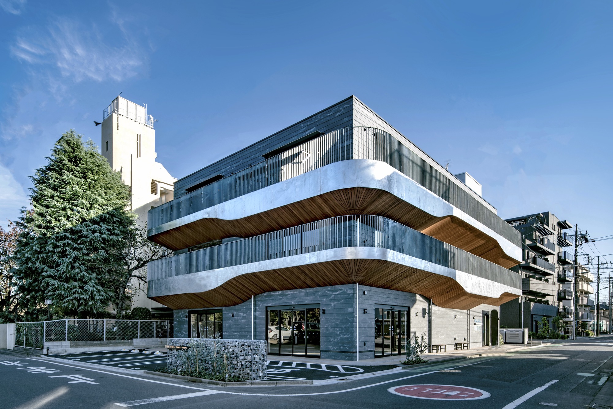 Street corner view of the layered facade with timber soffits and gabion walls under clear blue sky